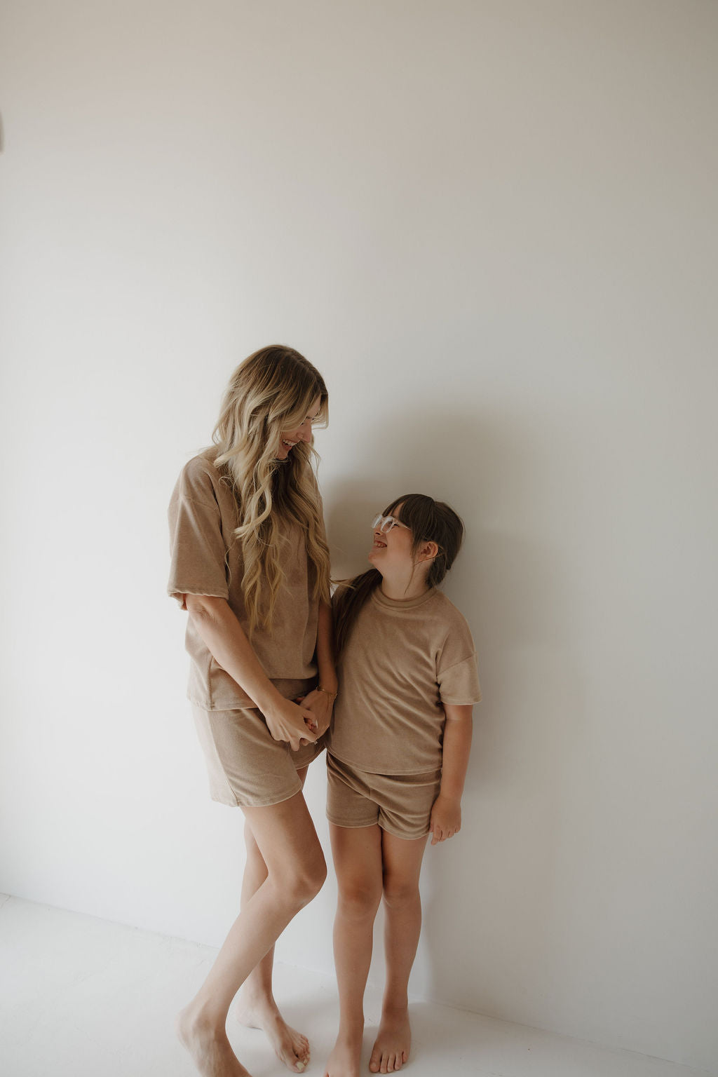 A woman and child stand barefoot against a white wall, smiling at each other as they hold hands. The child wears the forever french baby Child Terry Short Set in Tan, matching the woman's beige outfit for a warm family moment.