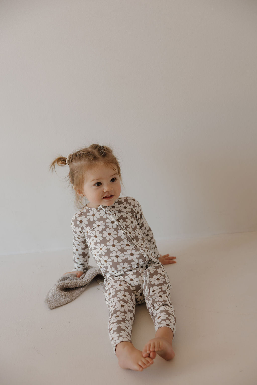 A young child with light brown pigtails sits barefoot, hugging a gray blanket and wearing forever french baby's Bamboo Zip Pajamas | Darlin', featuring a white daisy pattern on beige. The breathable bamboo fabric keeps her comfortable and relaxed.