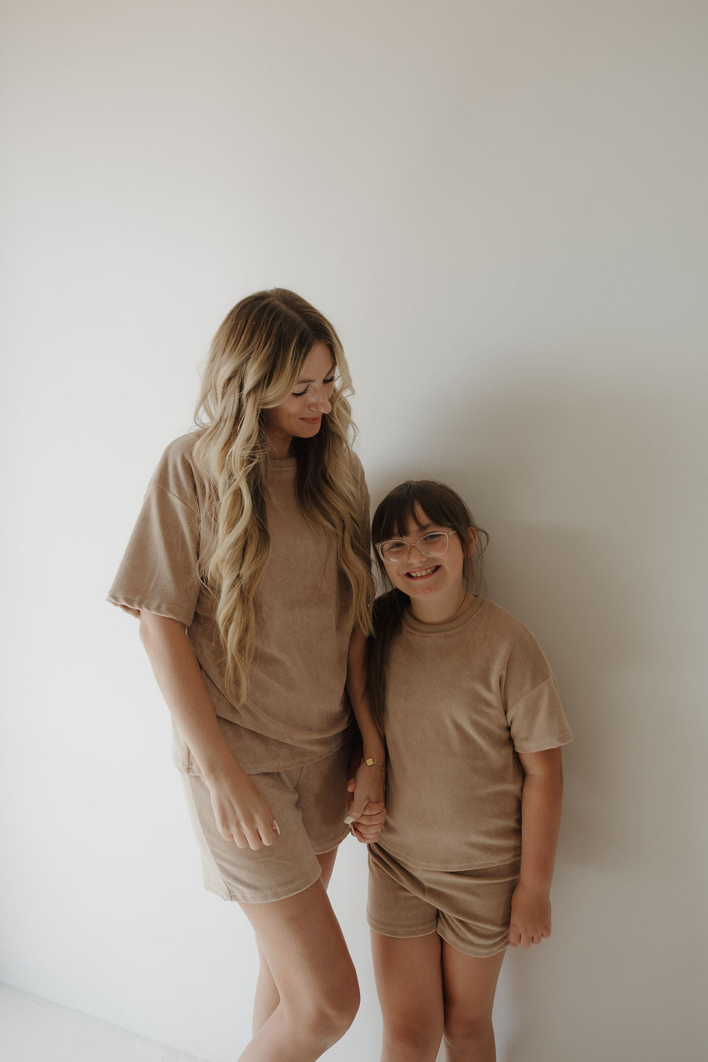 A woman and young girl stand against a white wall, both wearing the forever french baby Child Terry Short Set in tan. Smiling and holding hands, they capture a sweet family moment in matching outfits.