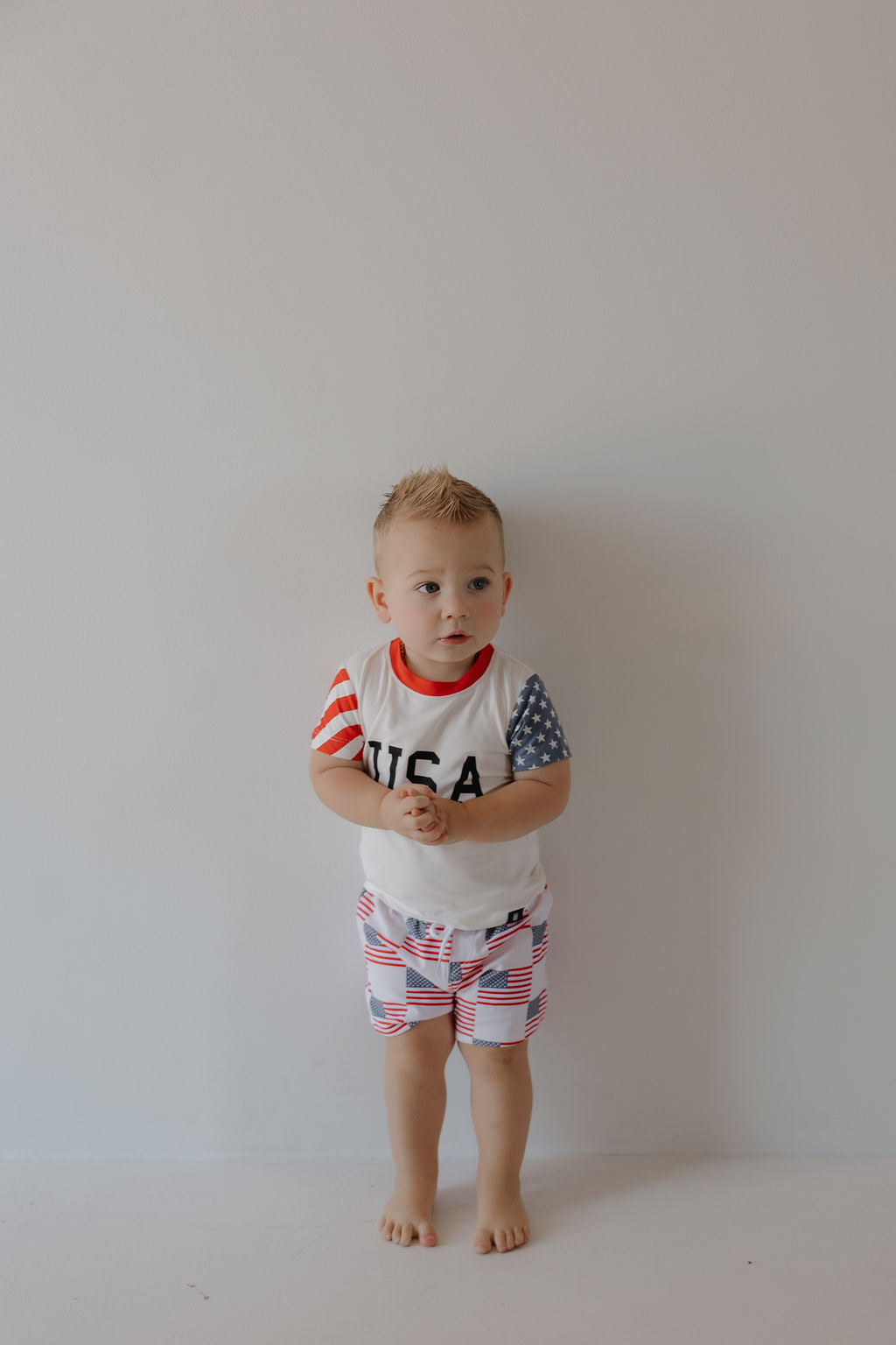 A young child with short blond hair stands barefoot against a plain white wall, wearing the forever french baby Child Bamboo T-Shirt | Stars & Stripes and matching shorts featuring USA flag-inspired designs.
