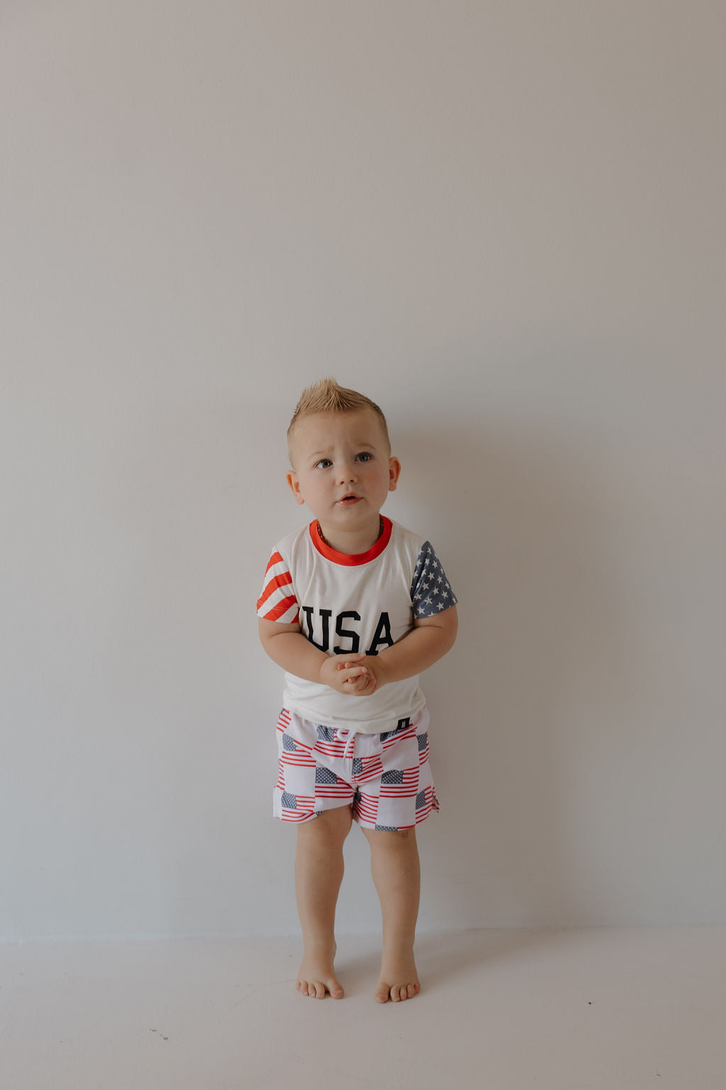 A young child with short blonde hair stands barefoot by a light wall, wearing the forever french baby Child Bamboo T-Shirt | Stars & Stripes and matching flag-patterned shorts made from breathable bamboo fabric.