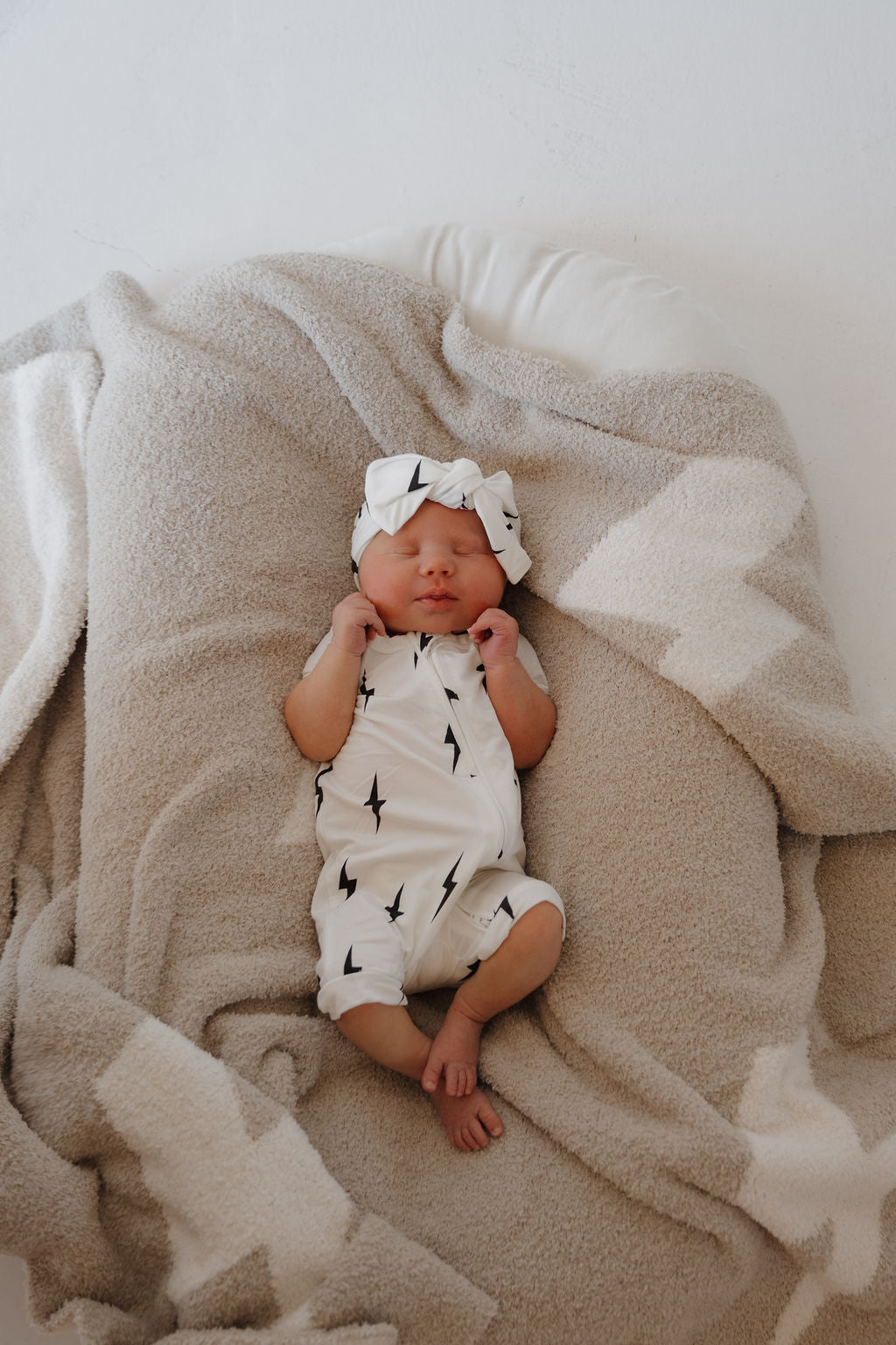 A newborn dressed in the forever french baby Bamboo Zip Shortie | White & Black Lightning Bolt with a matching headband sleeps on a soft beige and white blanket.