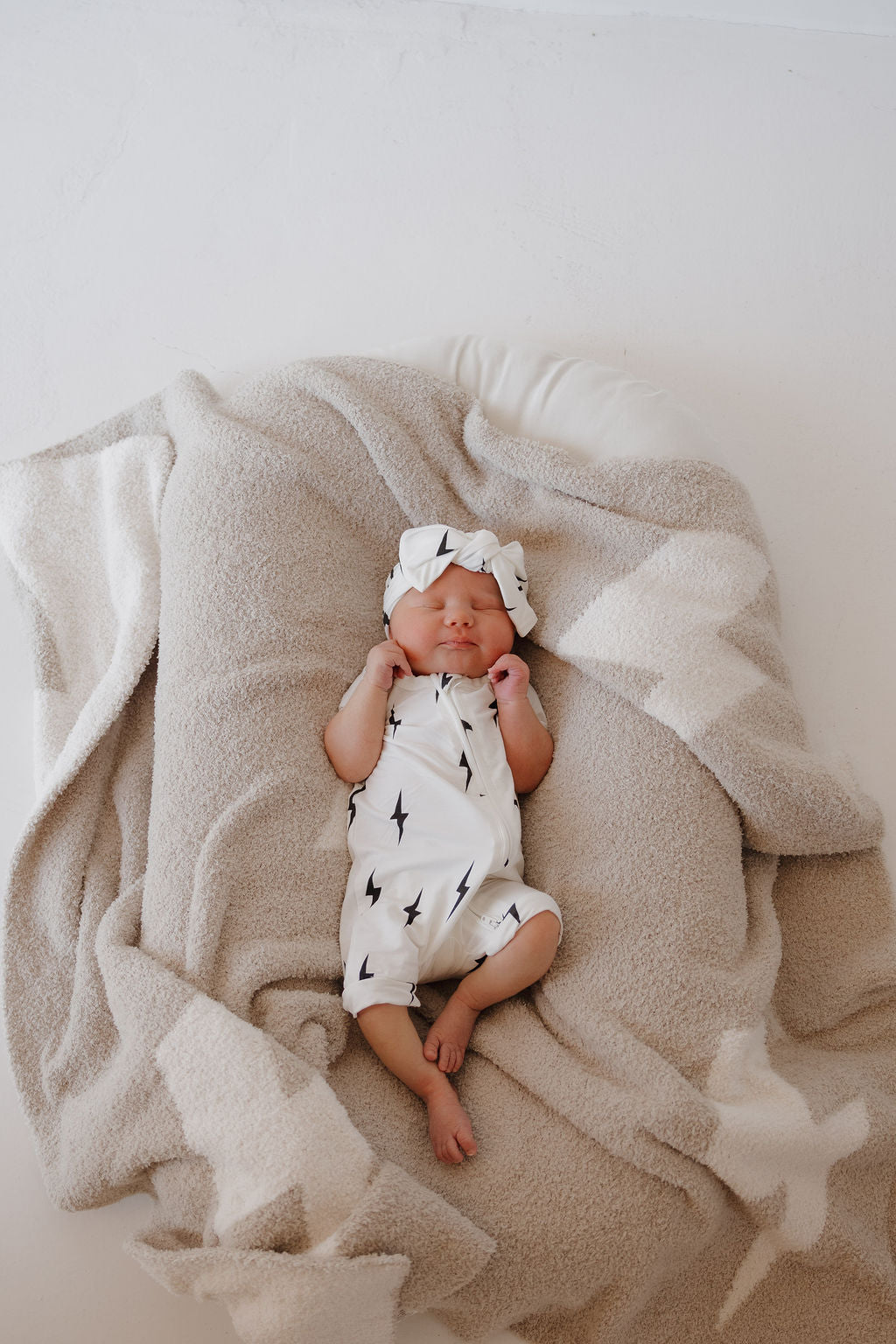 A newborn sleeps peacefully on a soft beige blanket, dressed in the forever french baby Bamboo Zip Shortie | White & Black Lightning Bolt and a matching headband.