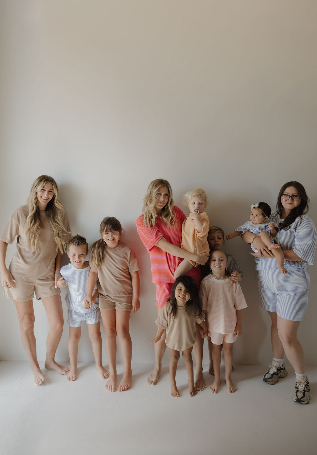 Three women and seven young children stand barefoot against a plain wall, relaxed and happy. Some of the little ones wear the forever french baby Child Terry Short Set in Ocean Stripe, dressed in casual, neutral-toned loungewear.