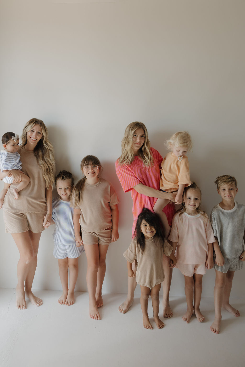A group of two women and seven children, including a toddler in the forever french baby Terry T-Shirt Romper | Ocean Stripe, stand barefoot against a light wall, wearing comfy pastel outfits—ideal for newborns to 24 months and easy diaper changes.