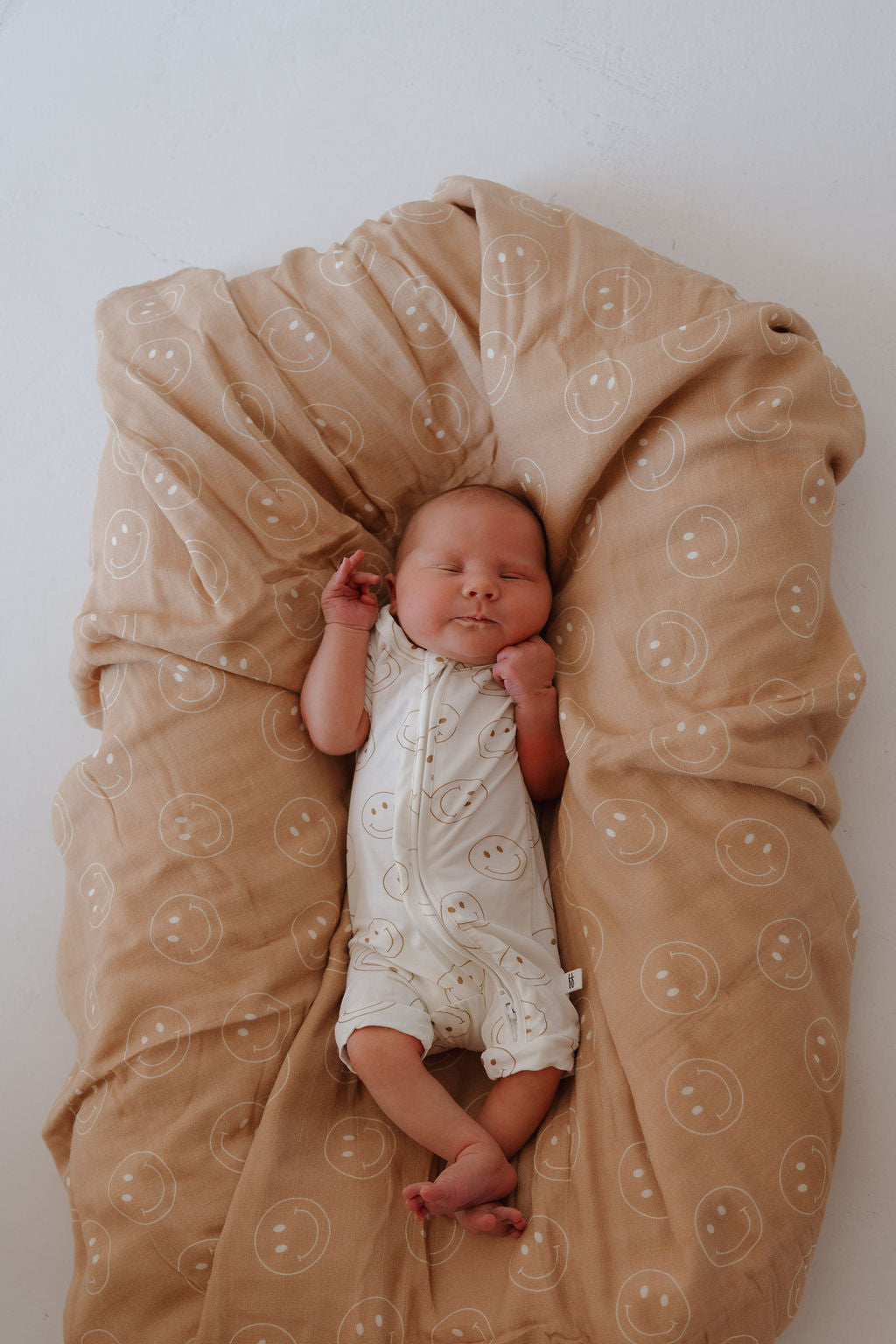 A newborn sleeps peacefully on a beige blanket with white smiley faces, wearing the forever french baby Bamboo Zip Shortie | Just Smile—breathable baby wear featuring a matching smiley face pattern.