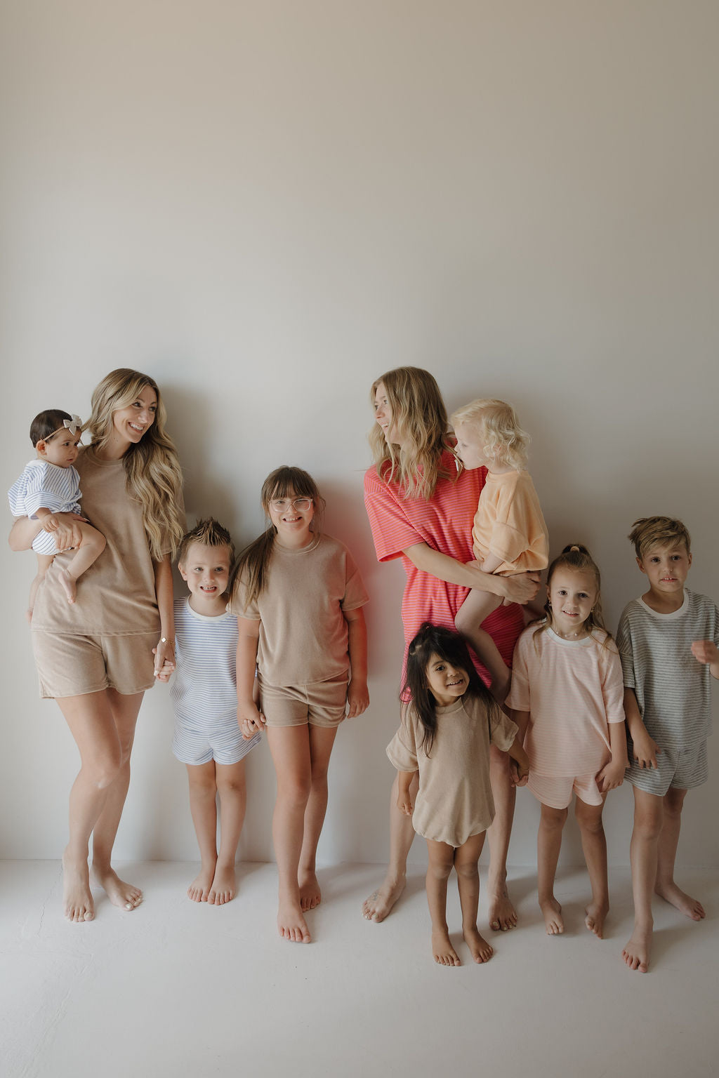 Two women stand against a plain wall, barefoot children in pastel loungewear around them. One cradles a baby in the forever french baby Terry T-Shirt Romper | Ocean Stripe—ideal for easy diaper changes from newborn to 24 months.