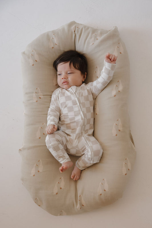 A dark-haired baby sleeps on a beige patterned cushion, wearing forever french baby Bamboo Zip Pajamas in Checkerboard—a light, breathable onesie—with one arm raised and eyes closed.