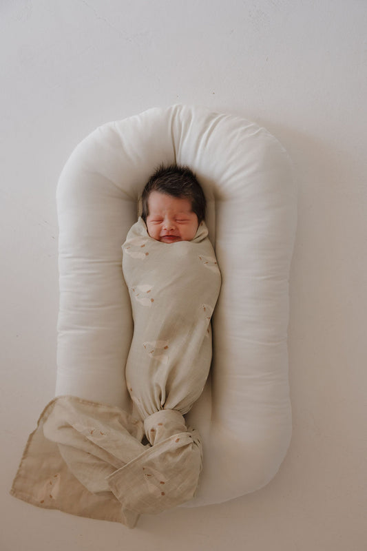 A newborn with dark hair sleeps peacefully on a white cushioned lounger, swaddled in a forever french baby Muslin Swaddle | Silly Goose, atop a light surface.