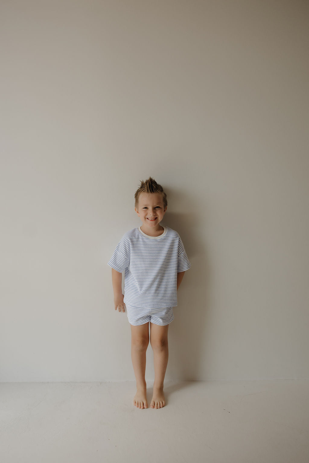 A young child with short, spiky hair stands barefoot against a plain light wall, smiling in the forever french baby Child Terry Short Set | Ocean Stripe—ideal for capturing sweet family moments.