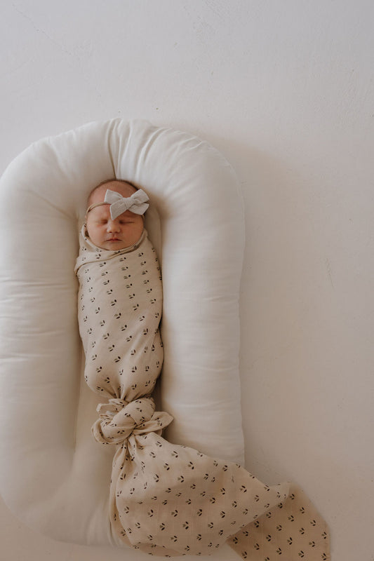 A newborn wrapped in the forever french baby Muslin Swaddle | Flora with a bow headband, resting on a soft white baby lounger against a plain light background.