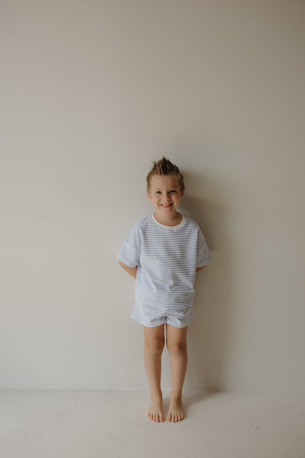 A young boy with short, spiky hair stands barefoot against a plain wall, smiling and wearing the forever french baby Child Terry Short Set | Ocean Stripe—an ideal outfit for capturing cherished family moments.
