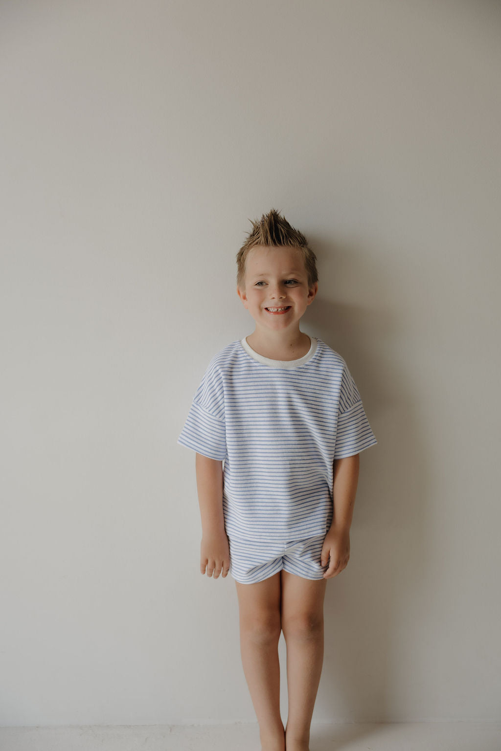 A young boy with short, spiky hair smiles barefoot against a white wall, wearing the forever french baby Child Terry Short Set | Ocean Stripe—light blue and white stripes perfect for cozy family moments.