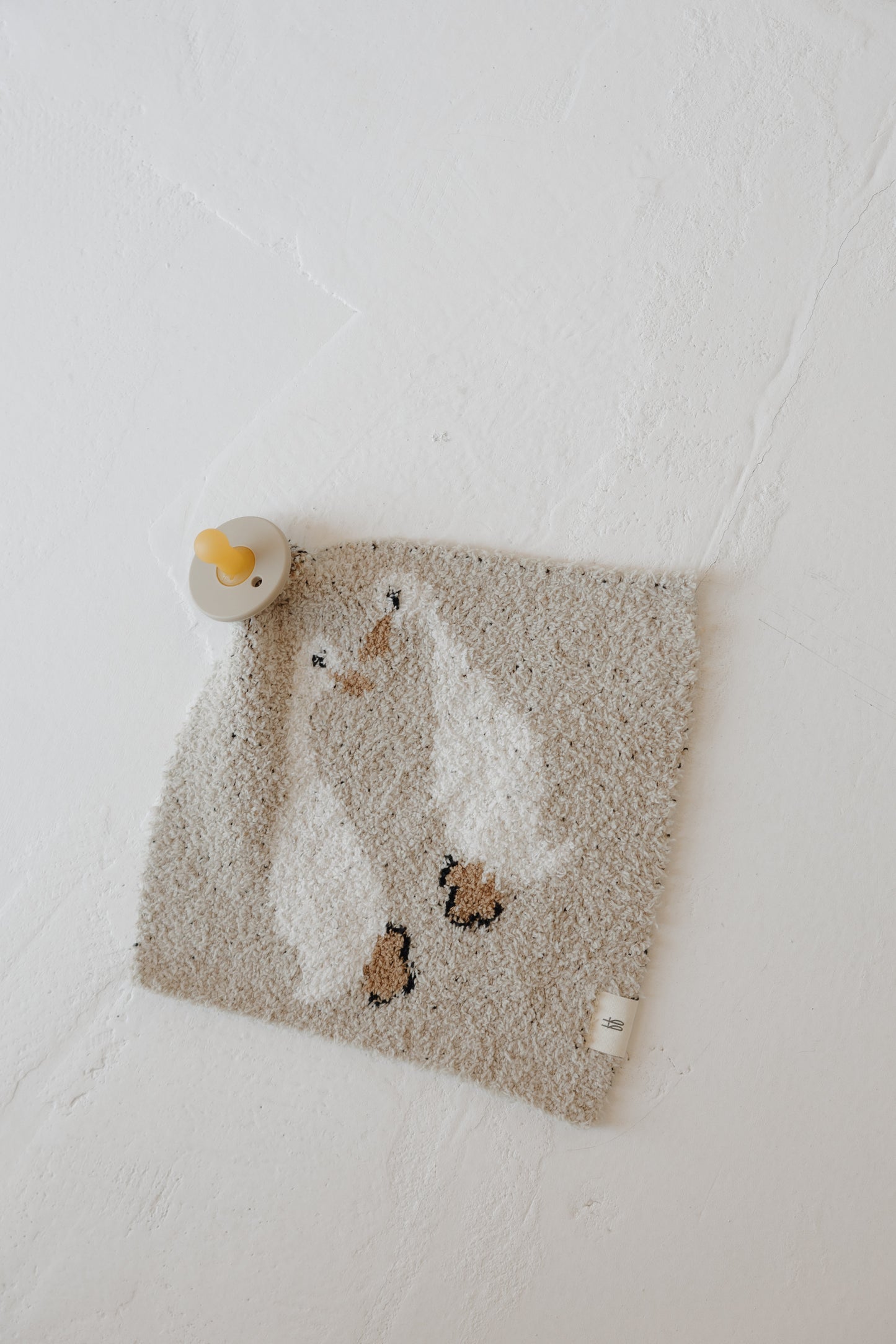 The Lovey | Silly Goose by forever french baby is a soft, square microfiber feather yarn washcloth featuring two white ducks. A yellow-and-gray pacifier rests on the upper left corner against a textured white background.