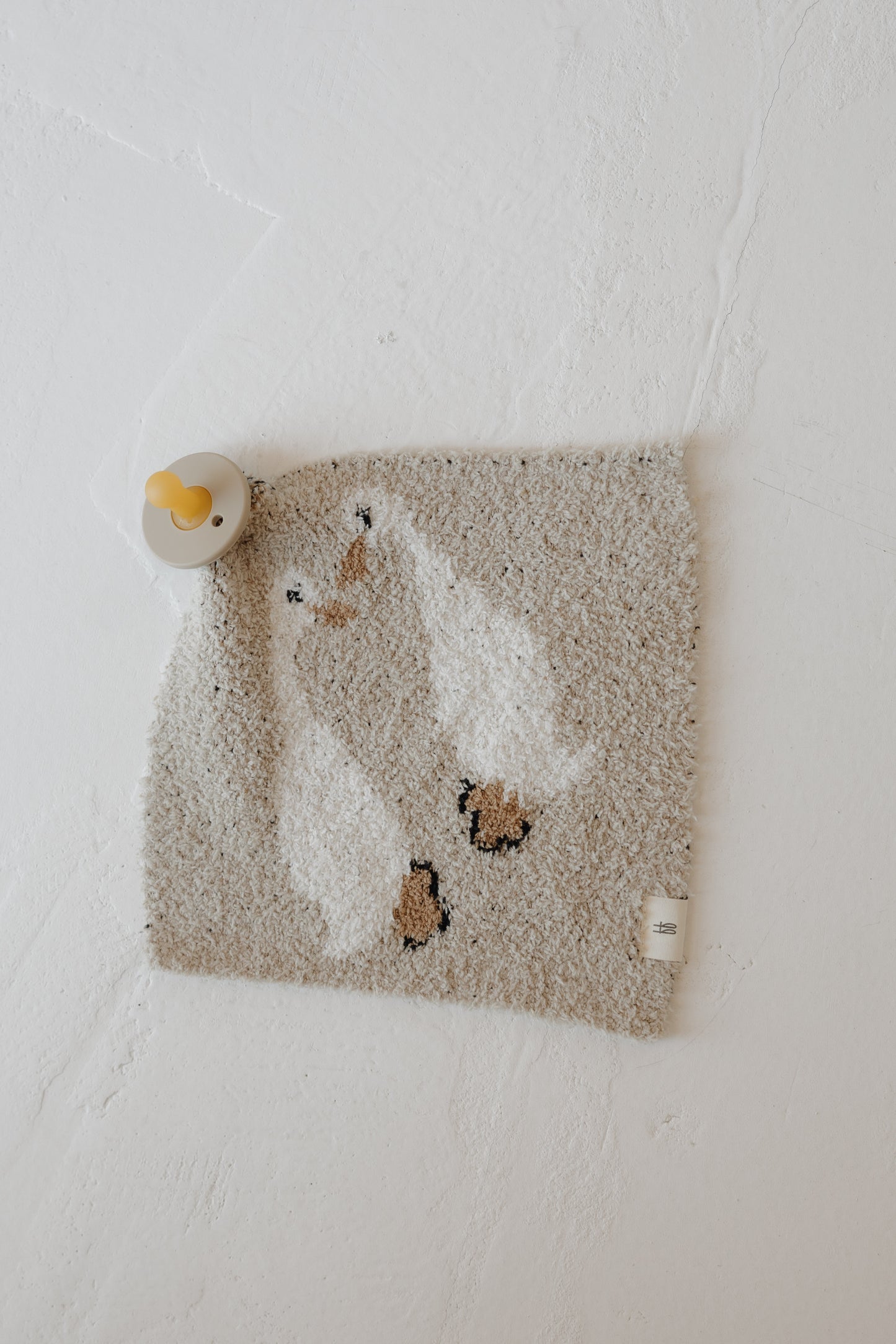 The Lovey | Silly Goose by forever french baby, a beige microfiber feather yarn towel with a white duck design, hangs on a yellow hook against a white wall.