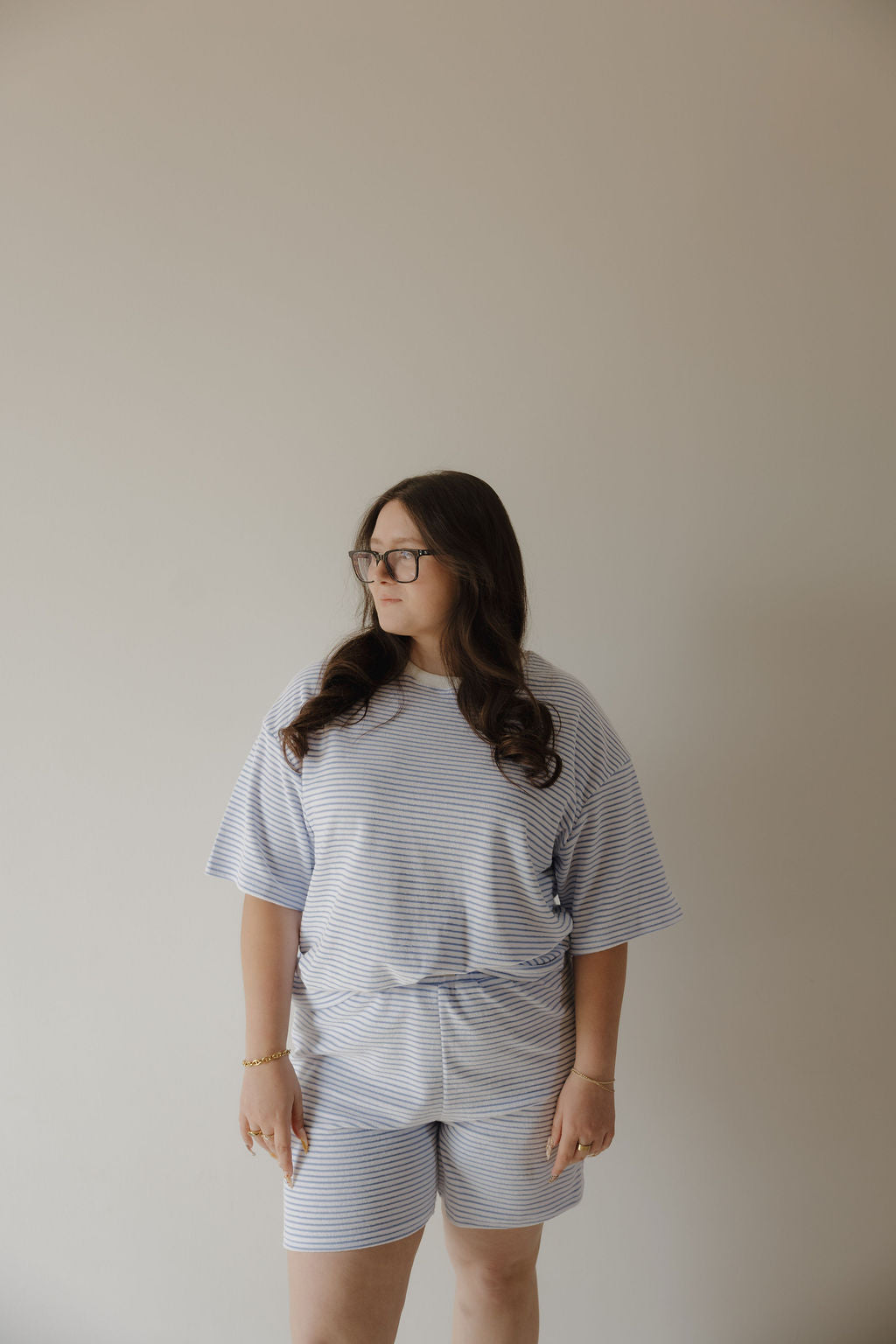 A woman with long dark hair and glasses stands against a plain light wall, wearing the Women's Terry Short Set in Ocean Stripe by forever french baby. She looks off to the side with a neutral expression.