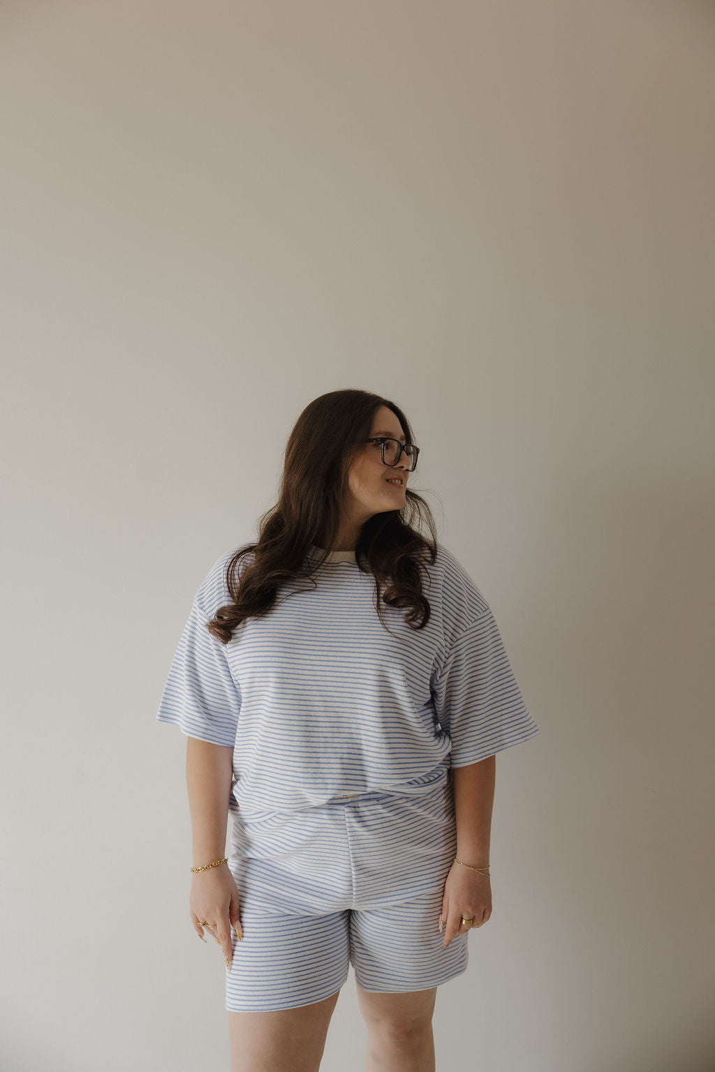 A woman with long brown hair and glasses stands against a plain light-colored wall, wearing the forever french baby Women's Terry Short Set in Ocean Stripe. She looks to the side with a relaxed expression.