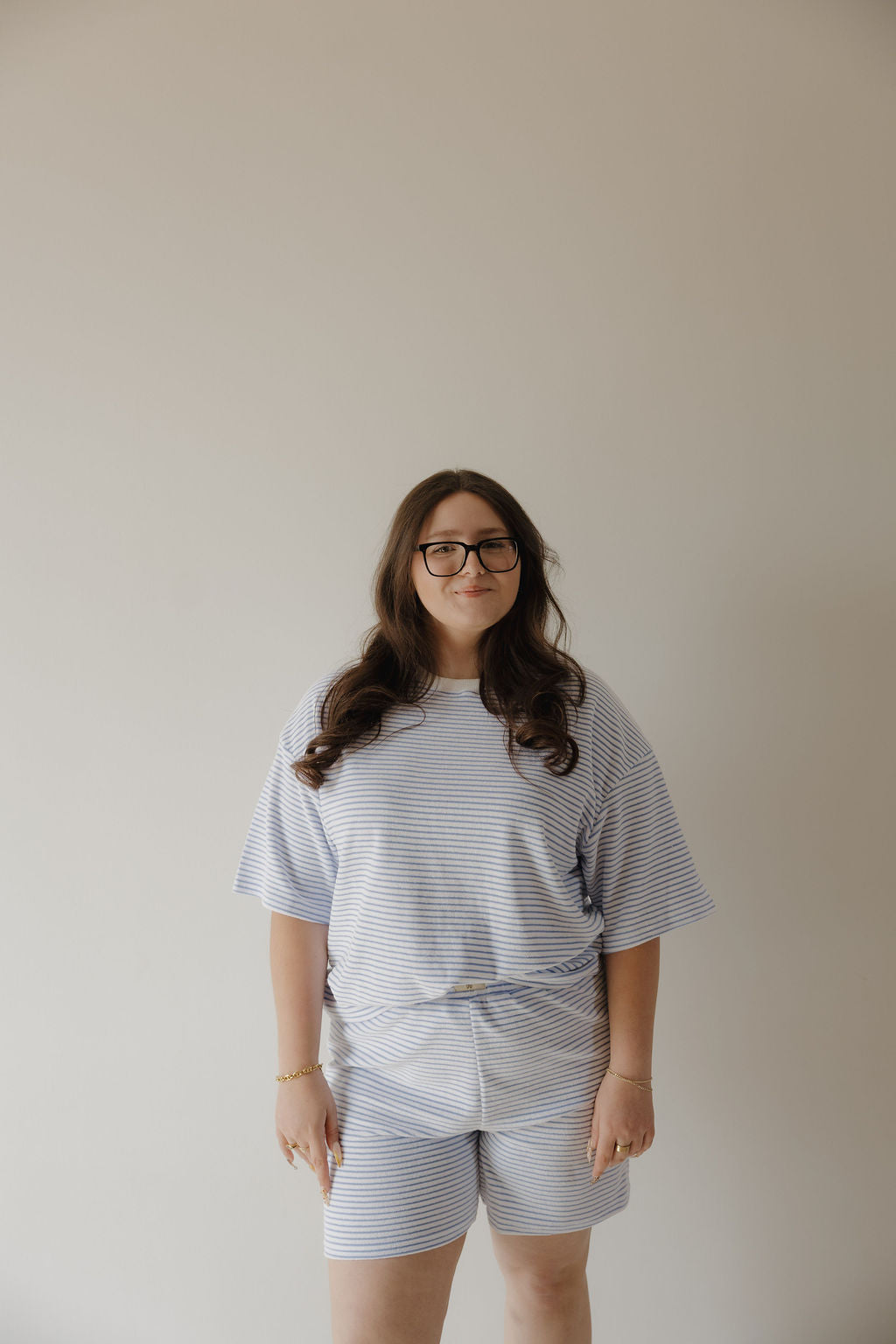 A person with long brown hair and glasses stands against a plain light-colored wall, wearing the Women's Terry Short Set in Ocean Stripe from forever french baby. They smile slightly with their hands relaxed by their sides.