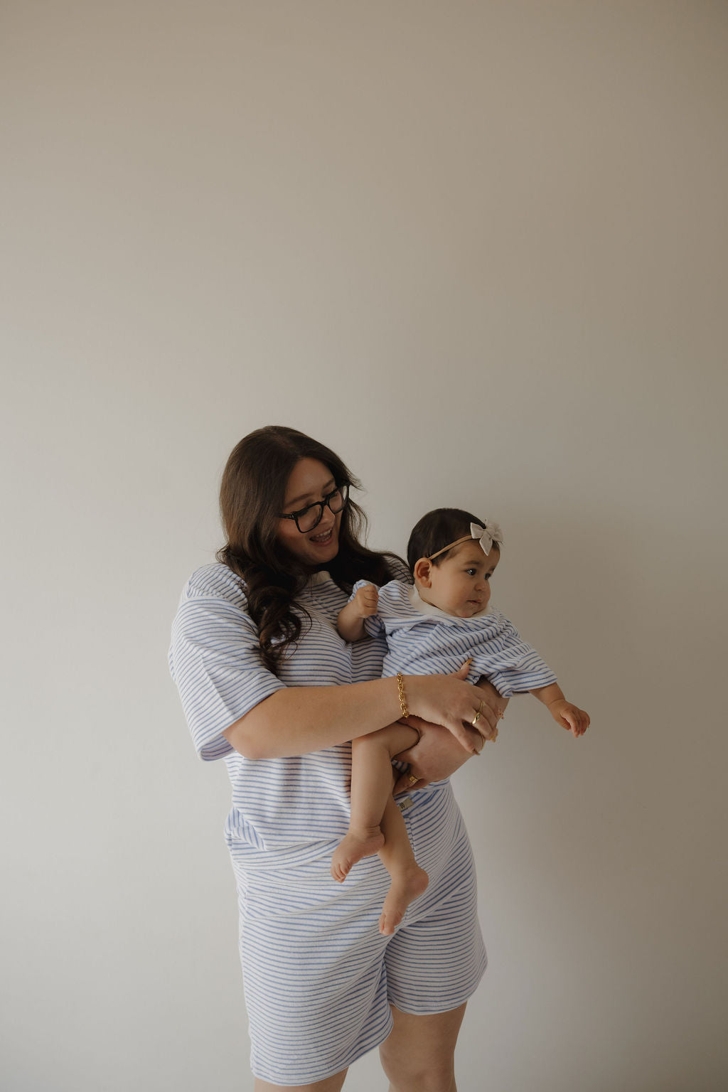 A woman with long brown hair and glasses holds a baby girl in matching forever french baby Terry T-Shirt Romper | Ocean Stripe, sized for newborn to 24 months. Both stand against a plain, light background, showcasing the easy-change outfit.