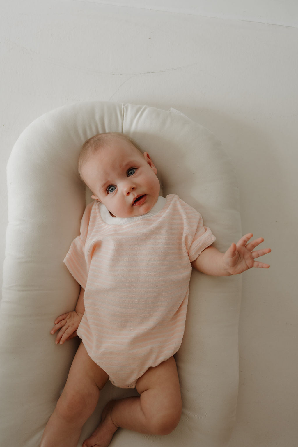 A baby wearing the forever french baby Terry T-Shirt Romper | Cotton Candy lies on a soft white cushion, looking up with one arm raised. Snap closures ensure comfort, and the minimal background highlights sweet family moments.