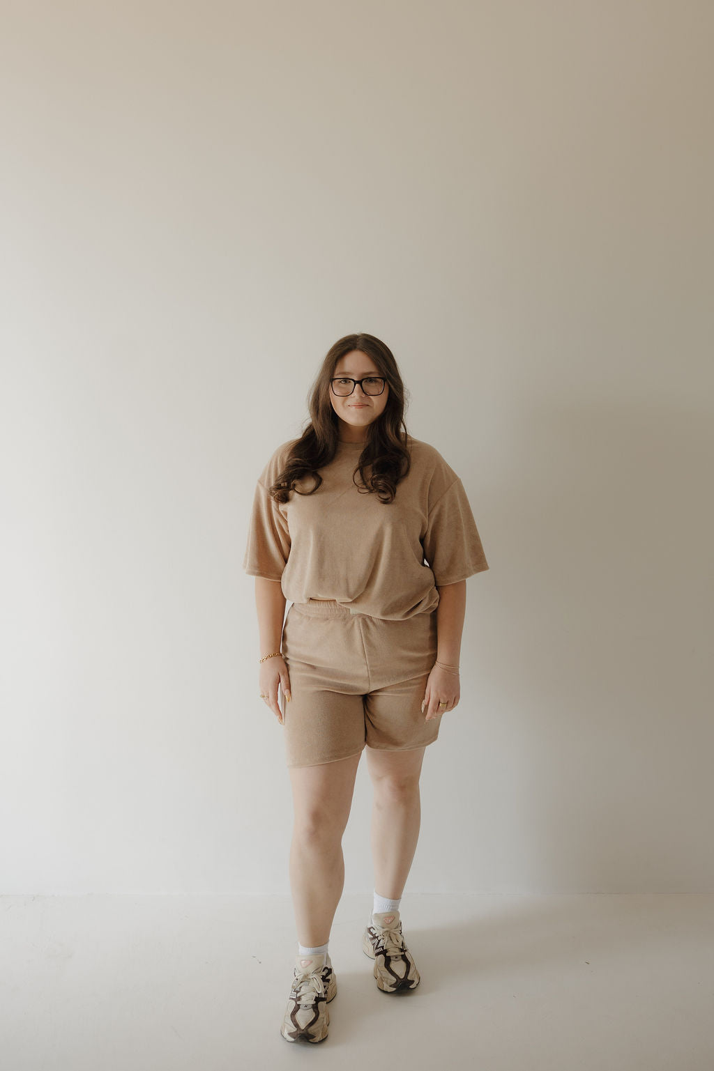 A woman with long brown hair and glasses stands against a plain wall, wearing the forever french baby Women's Terry Short Set in tan, paired with matching shorts, white socks, and sneakers.