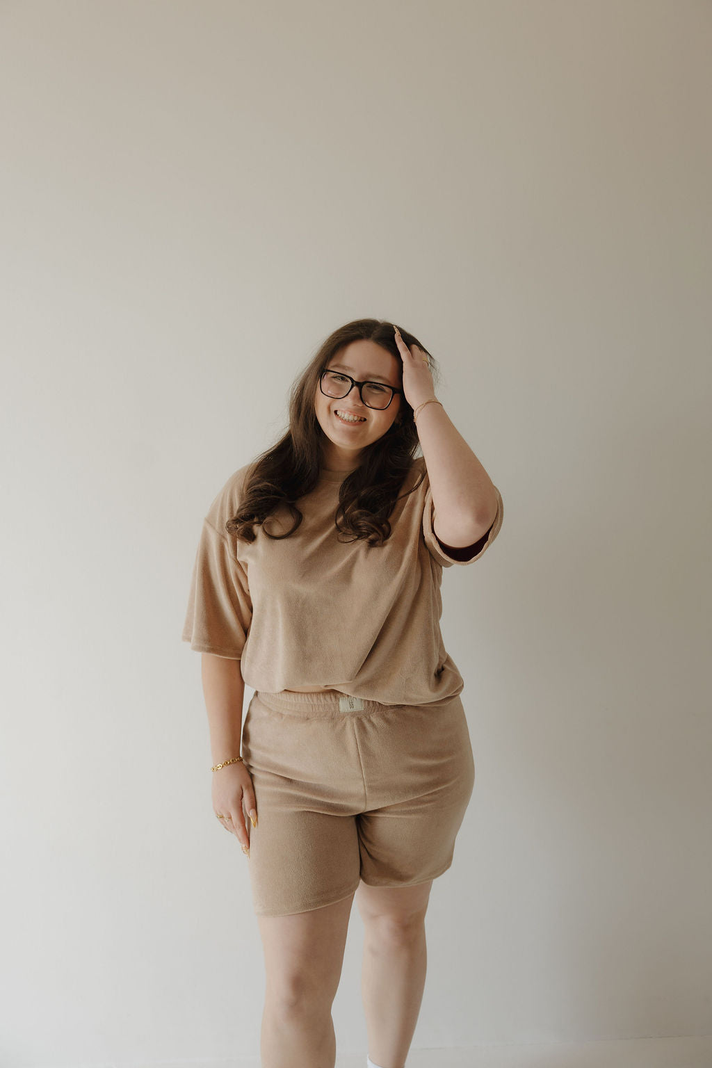 A woman with long brown hair and glasses stands against a light-colored wall, smiling as she touches her hair. She is wearing the forever french baby Women's Terry Short Set in tan—a matching T-shirt and shorts.