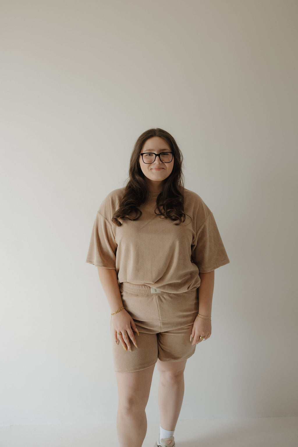 A woman with long brown hair and glasses smiles gently, standing against a light wall in the forever french baby Women's Terry Short Set in Tan—matching shorts and t-shirt—accessorized with bracelets and rings.