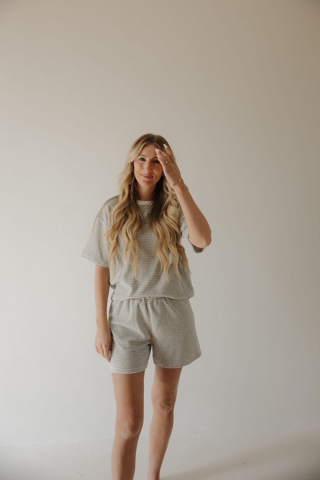 A woman with long, wavy blonde hair smiles softly, wearing the forever french baby Women's Terry Short Set in Grey Stripe against a beige wall—timeless comfort and style.