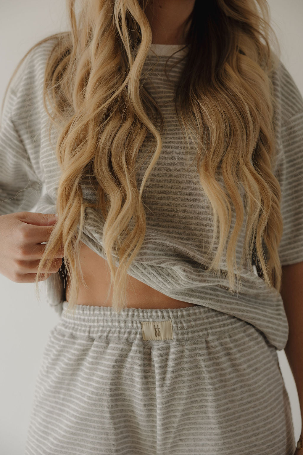 A woman with long wavy blonde hair wears the forever french baby Women's Terry Short Set in Grey Stripe, lifting her shirt slightly to show her midriff. The image is cropped from shoulders to thighs.