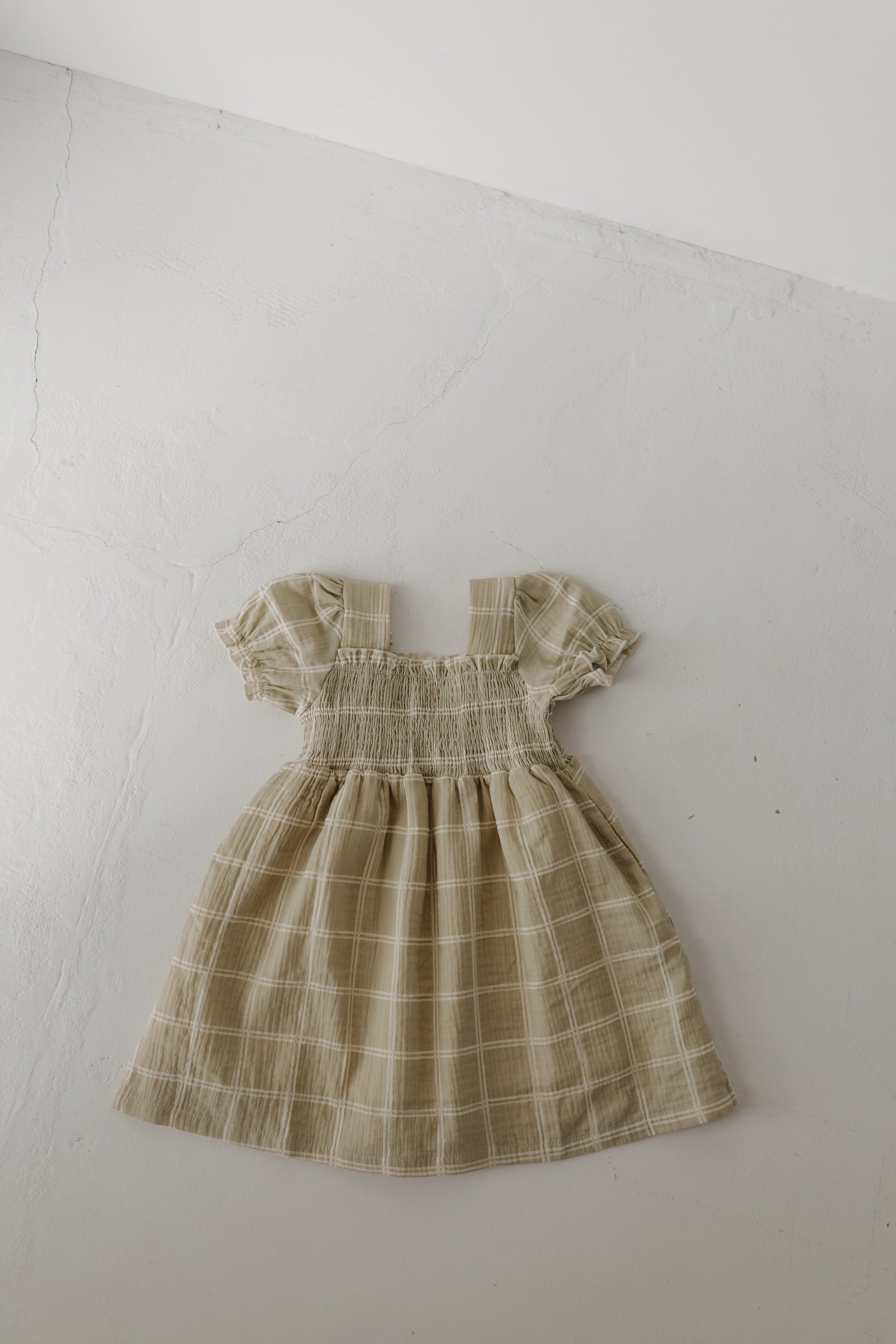 The Toddler Muslin Dress | Linen Grid by forever french baby is a beige plaid, short-sleeve 100% cotton dress with a smocked bodice and gathered skirt, shown laid flat on a light textured surface. Soft, breathable, and classic in design.