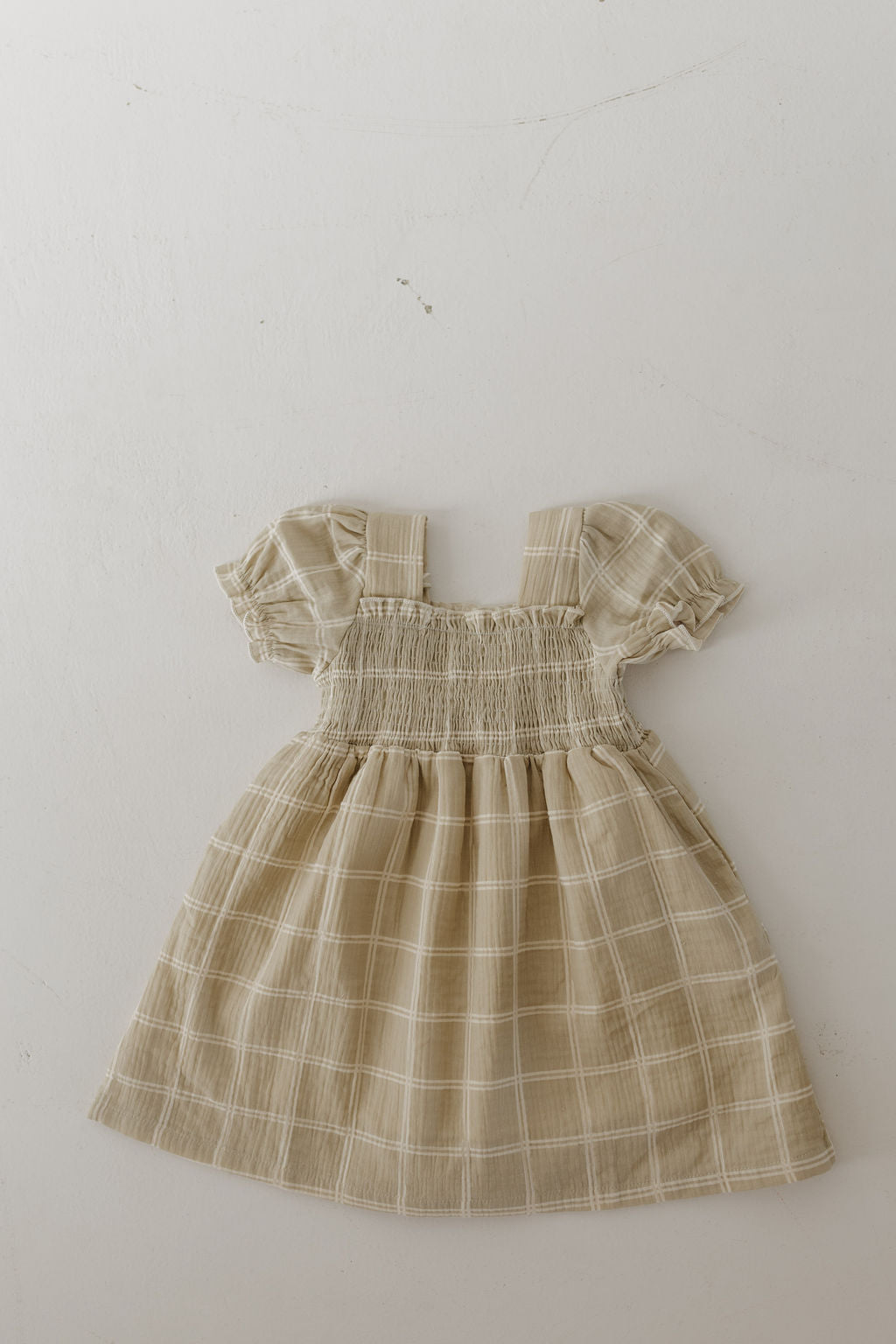The forever french baby Toddler Muslin Dress | Linen Grid features a beige plaid design with short puffed sleeves, smocked bodice, and gathered skirt in breathable 100% cotton muslin, shown laid flat on a light background.