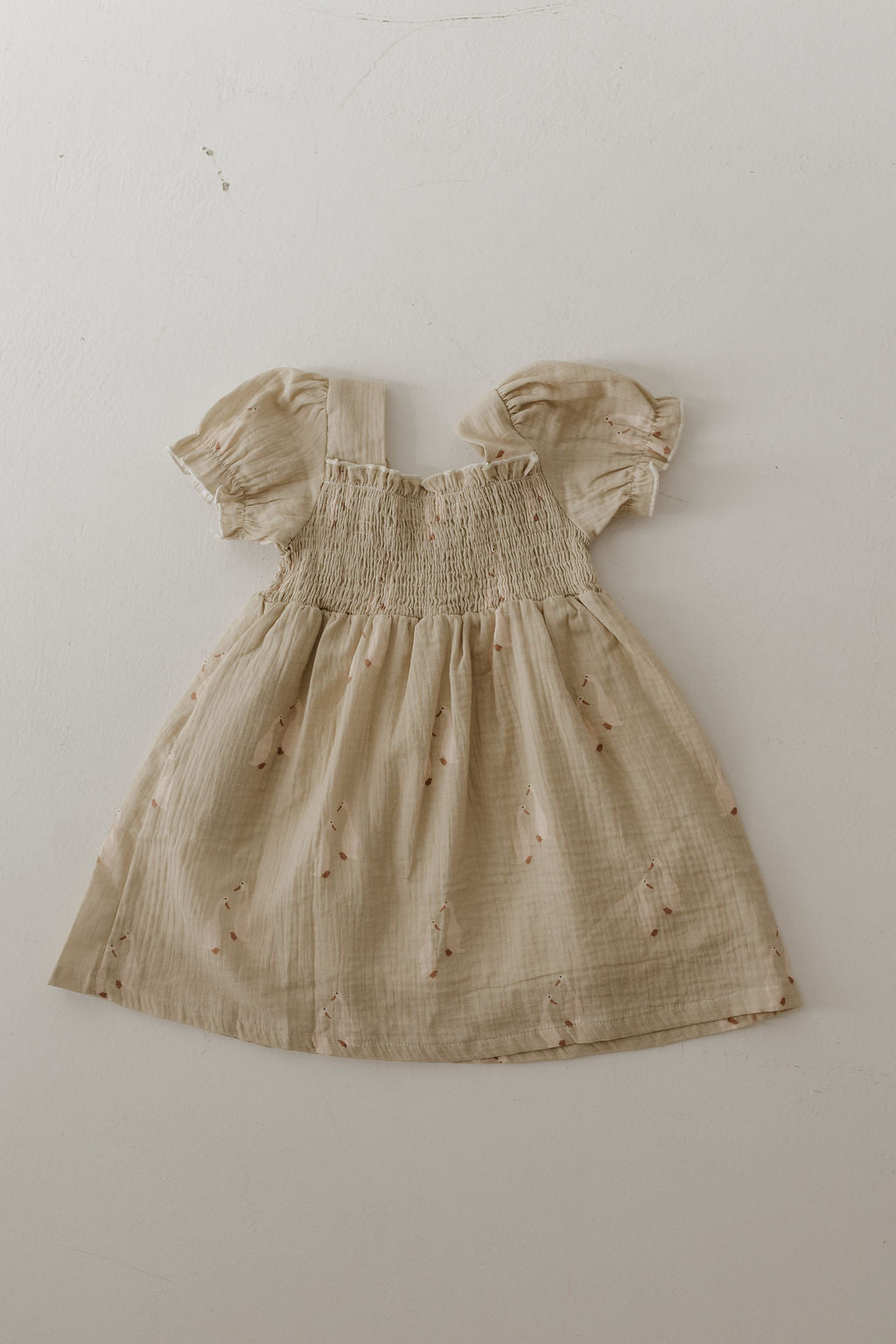 The forever french baby Toddler Muslin Dress | Silly Goose is a beige, short-sleeved dress with puff sleeves, smocked bodice, gathered skirt, and delicate floral prints. Made from 100% cotton muslin with a subtle textured pattern. Laid flat on a light surface.