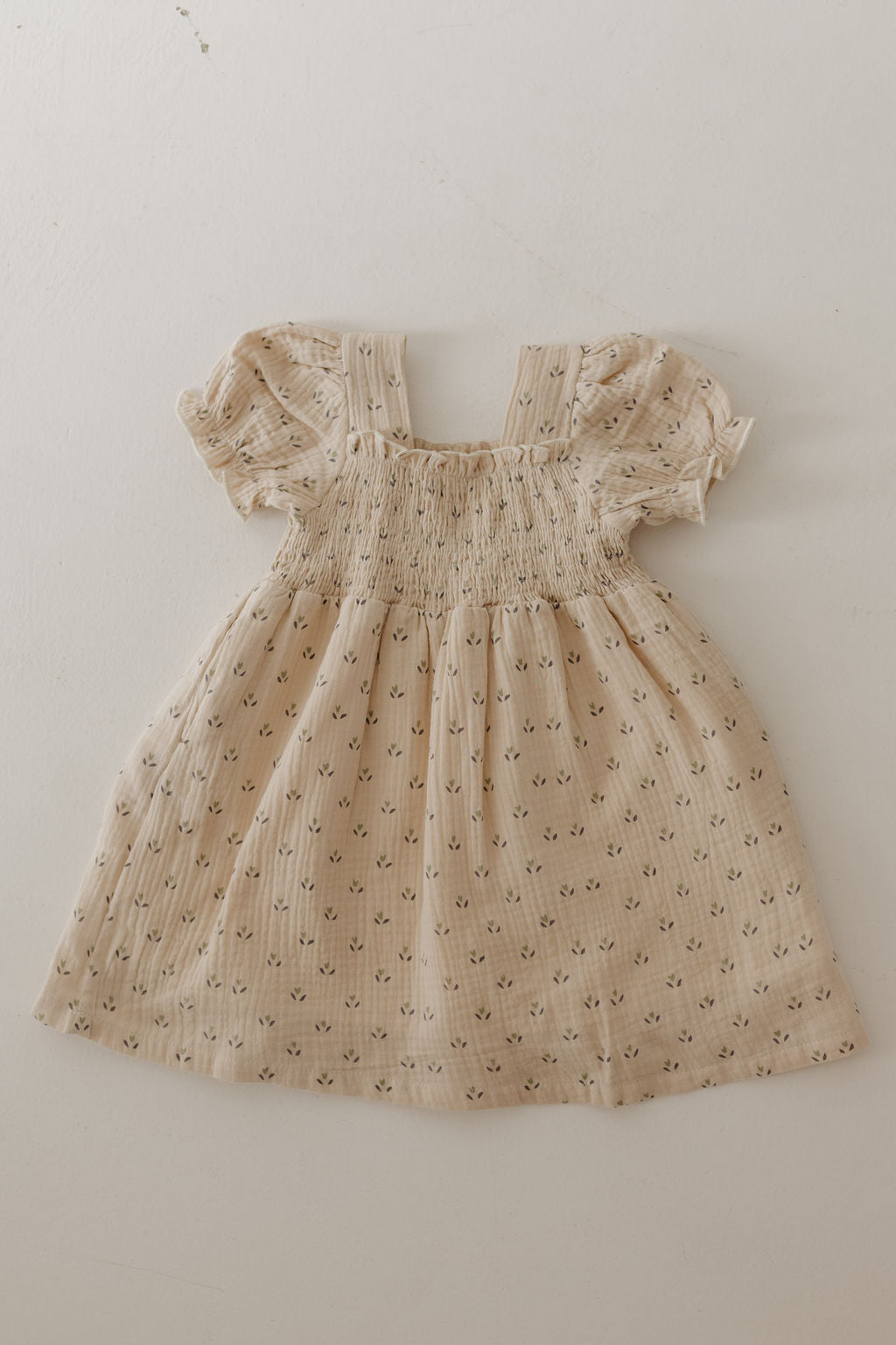 The Toddler Muslin Dress | Flora by forever french baby is a light beige, short-sleeve 100% cotton dress for babies with a smocked bodice and small floral print—perfect for cherished mom-and-me moments. Laid flat on a white surface.