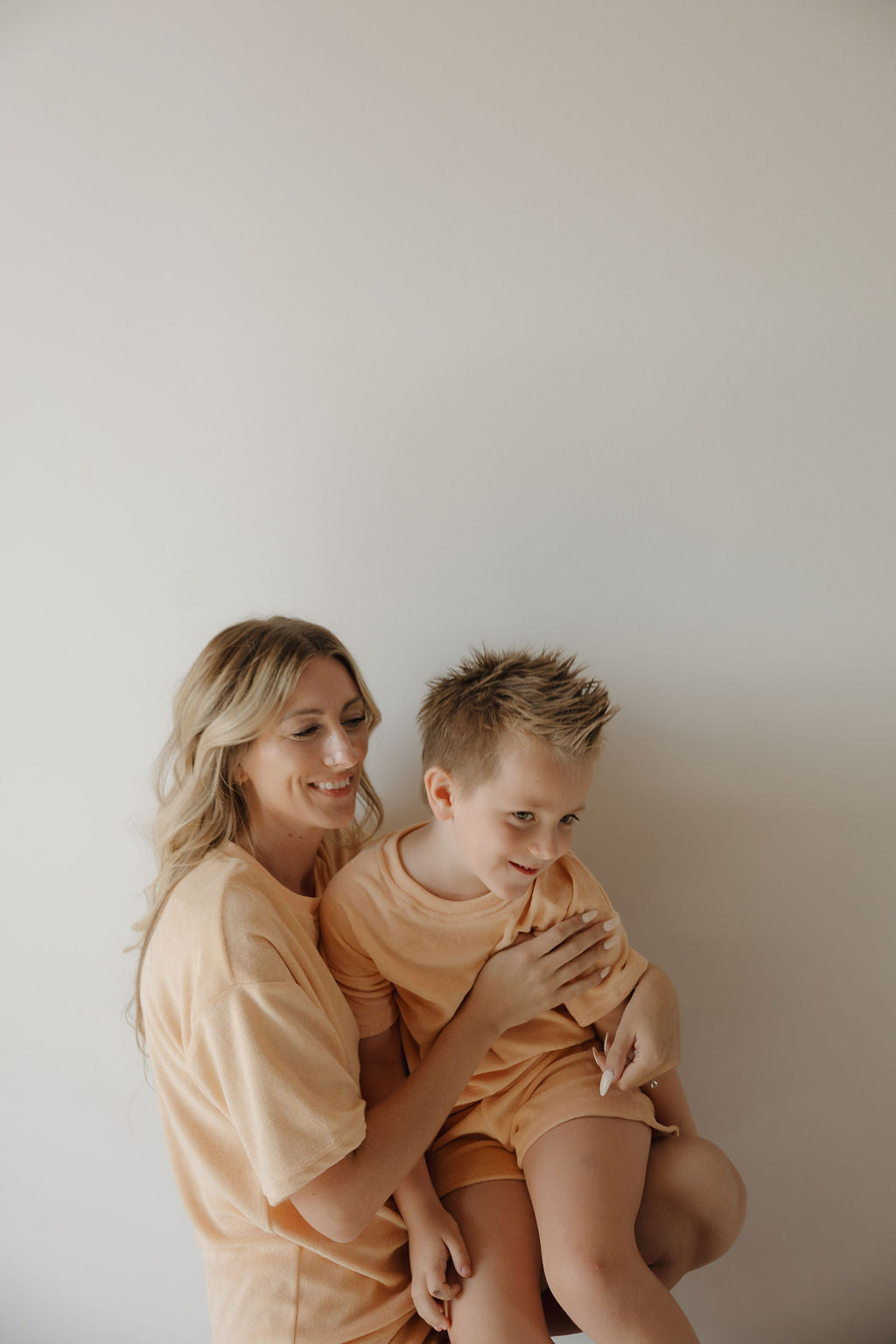 A smiling woman with long blonde hair hugs a young boy, both wearing matching forever french baby Chid Terry Short Set in Sherbert, as they sit together against a plain white background—capturing heartfelt family moments.