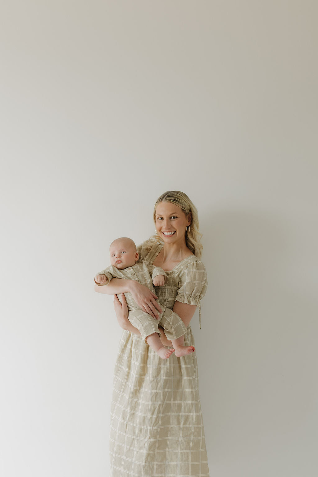A woman smiles while holding a baby in matching outfits from forever french baby: she wears the Women's Short Sleeve Muslin Dress | Linen Grid, and the baby is dressed in a coordinating 100% cotton set.