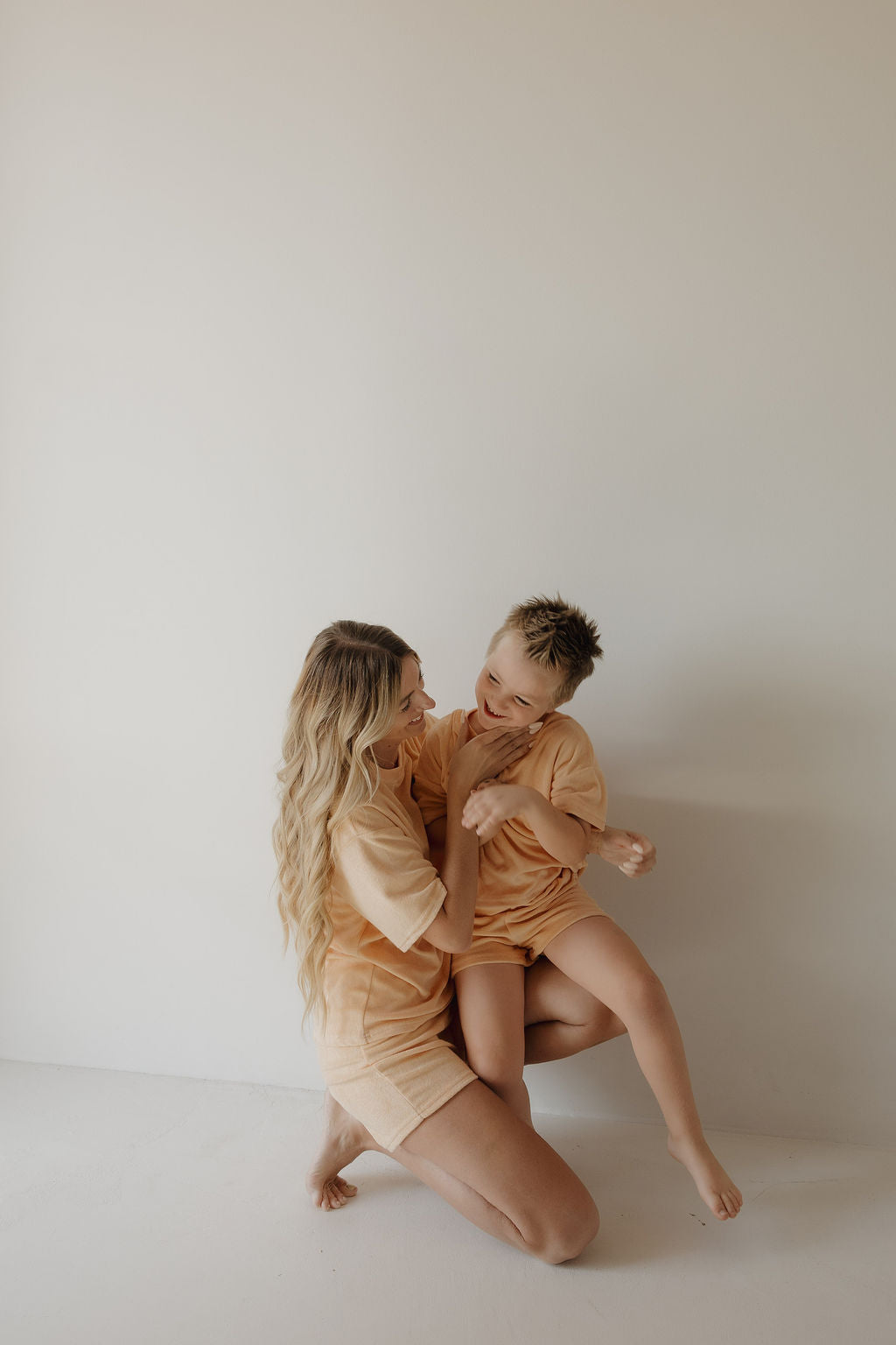 A woman with long, wavy blonde hair kneels on the floor, smiling and playing with a laughing boy. Both wear matching forever french baby Child Terry Short Set | Sherbert, capturing joyful family moments against a plain, light background.