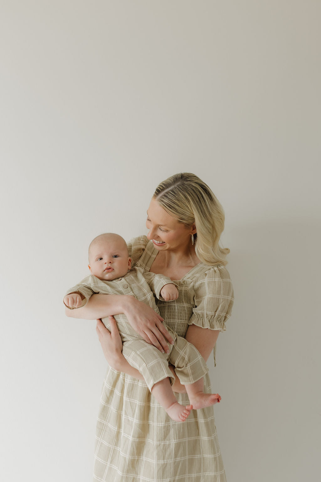 A smiling woman in the forever french baby Women's Short Sleeve Muslin Dress in Linen Grid holds a baby in matching plaid outfits against a plain light background.