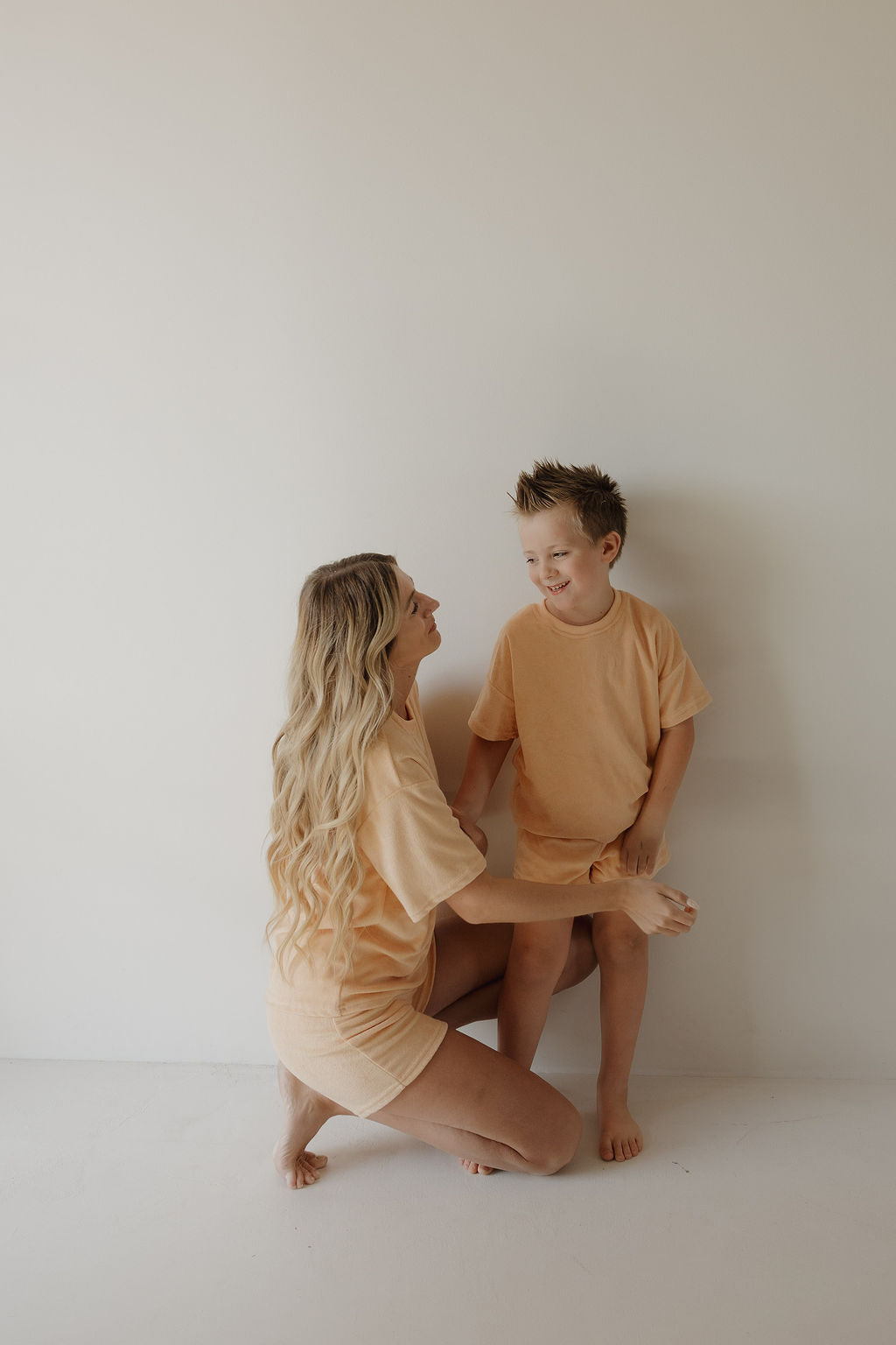 A woman with long blonde hair kneels and smiles at a young boy. Both wear matching forever french baby loungewear, including the Child Terry Short Set | Sherbert, against a plain wall—a minimalistic look perfect for family moments.