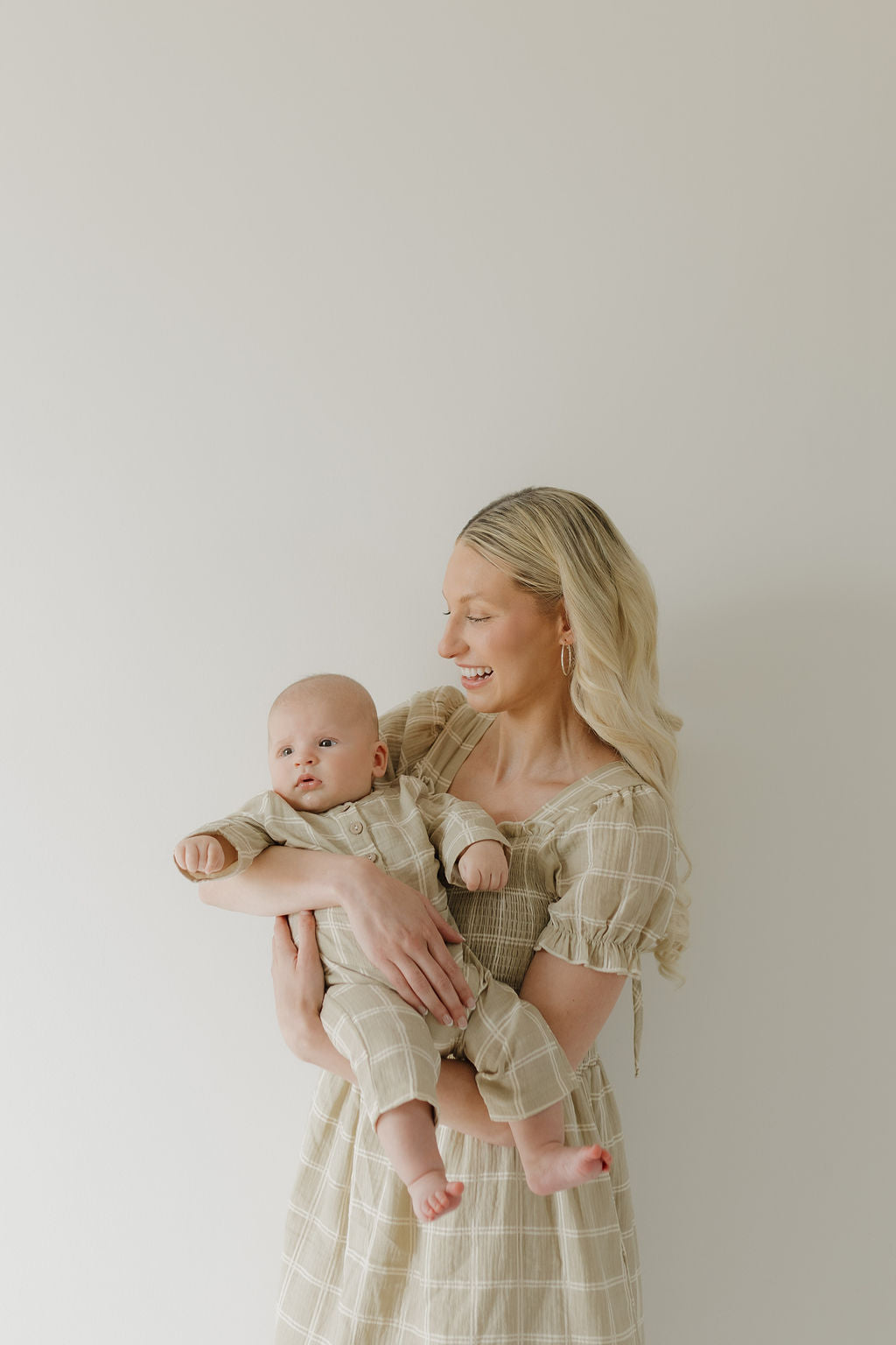 A woman with long blonde hair wears the forever french baby Women's Short Sleeve Muslin Dress in Linen Grid as she holds a baby in matching outfits. Both smile and look to the side against a plain, light-colored background.