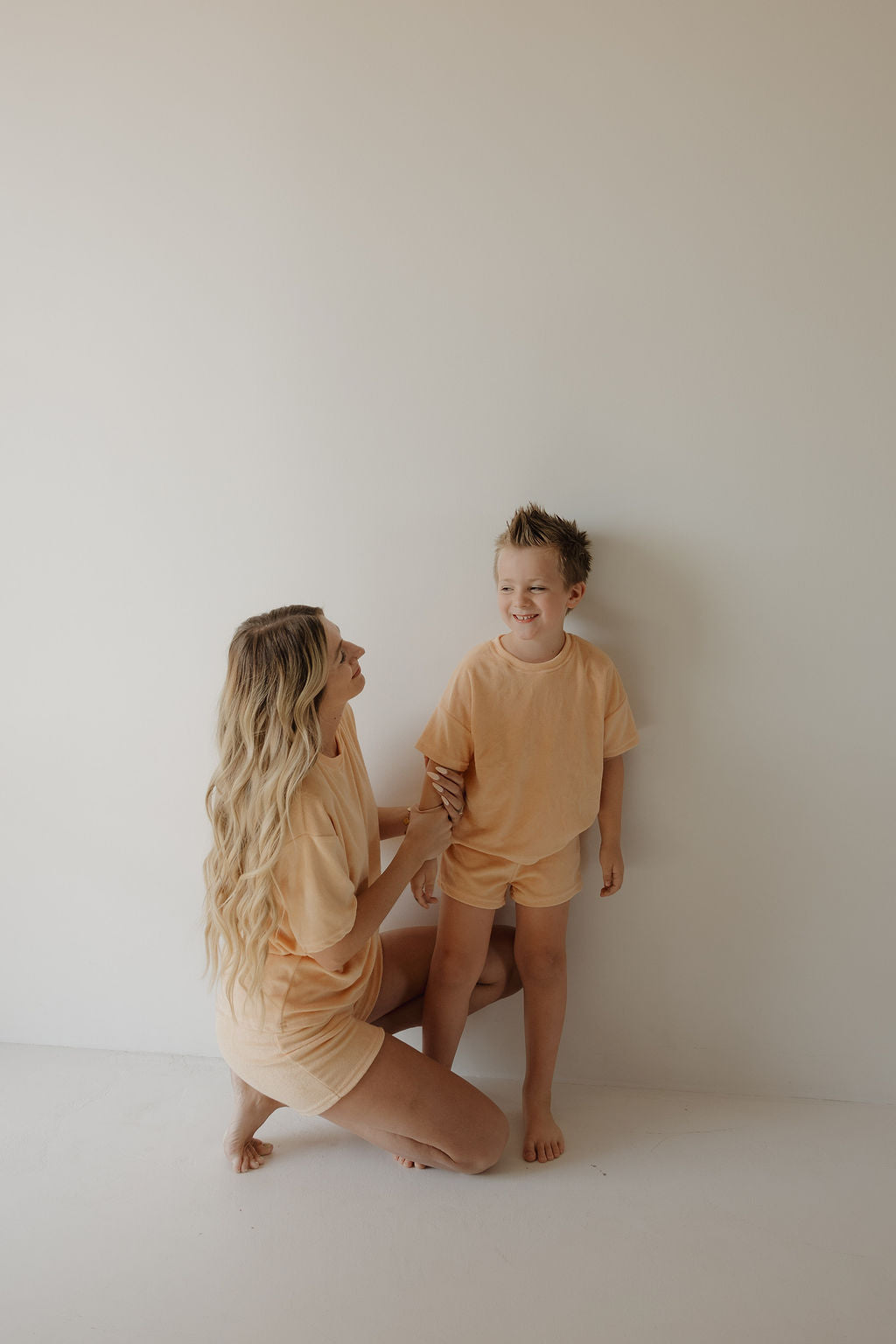 A woman with long blonde hair kneels beside a smiling young boy, both wearing matching Sherbert Chid Terry Short Sets from forever french baby, radiating joy against a plain light background and capturing bright, minimalistic family moments.