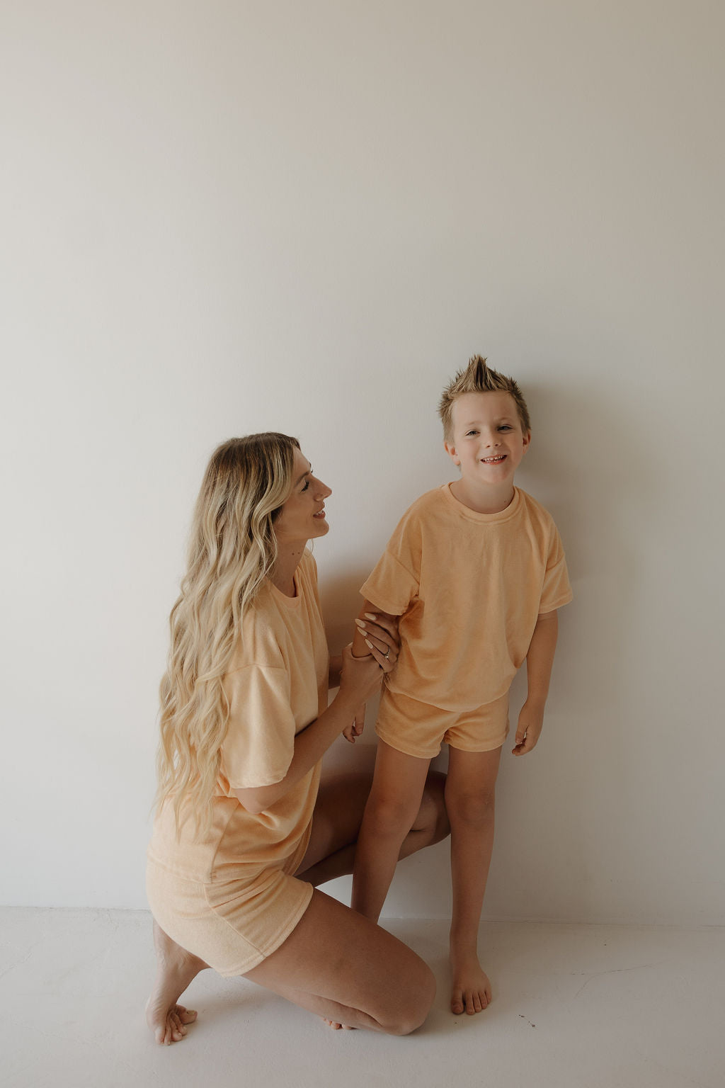 A woman kneels beside a smiling young boy in the forever french baby Chid Terry Short Set | Sherbert; both wear matching peach loungewear, sharing family moments against a light wall as she gently holds his arm and looks up at him.