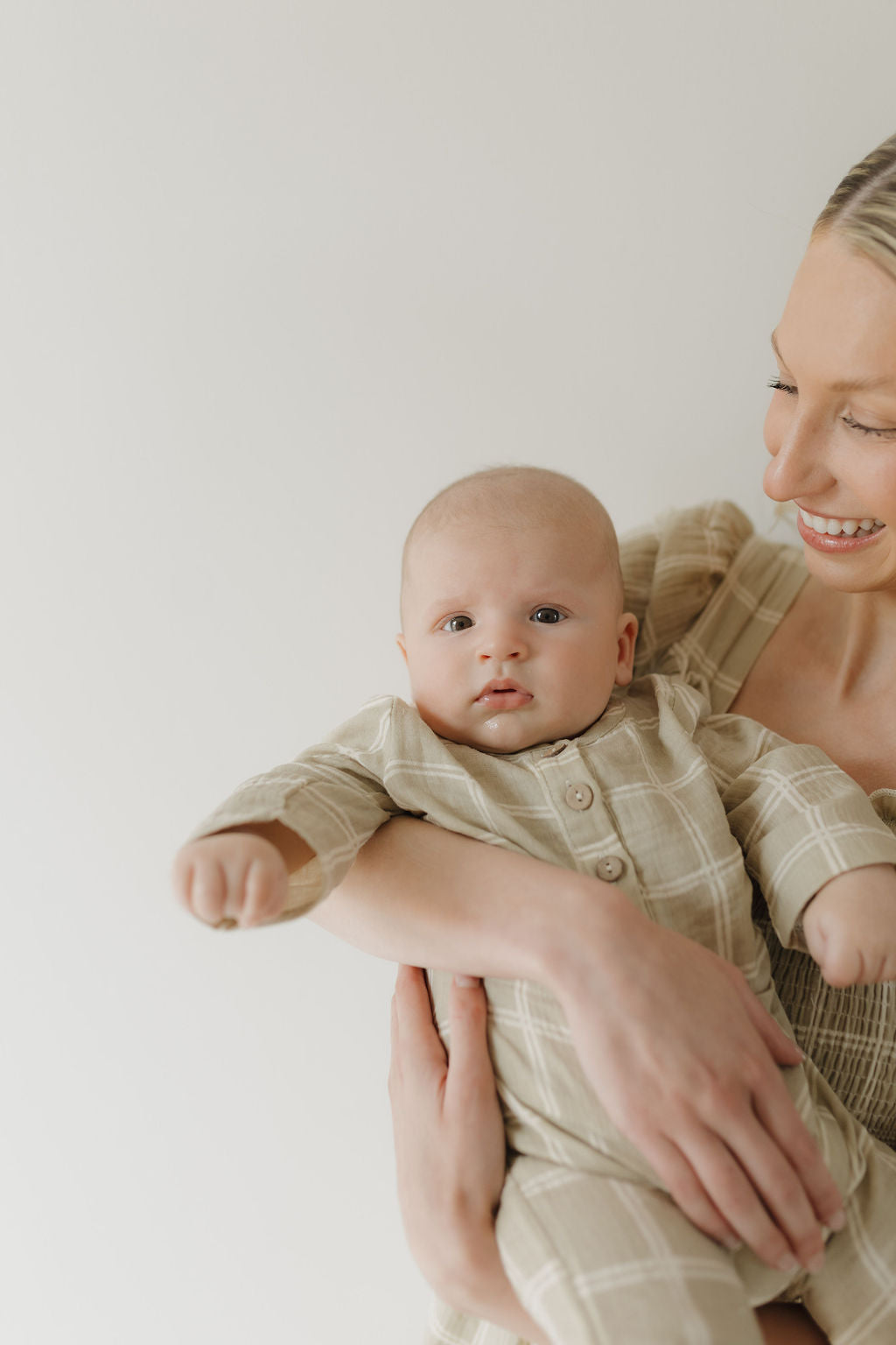 A smiling woman wears the forever french baby Women's Short Sleeve Muslin Dress in Linen Grid while holding a baby. The baby, dressed in beige plaid, reaches toward the camera against a light, airy background.