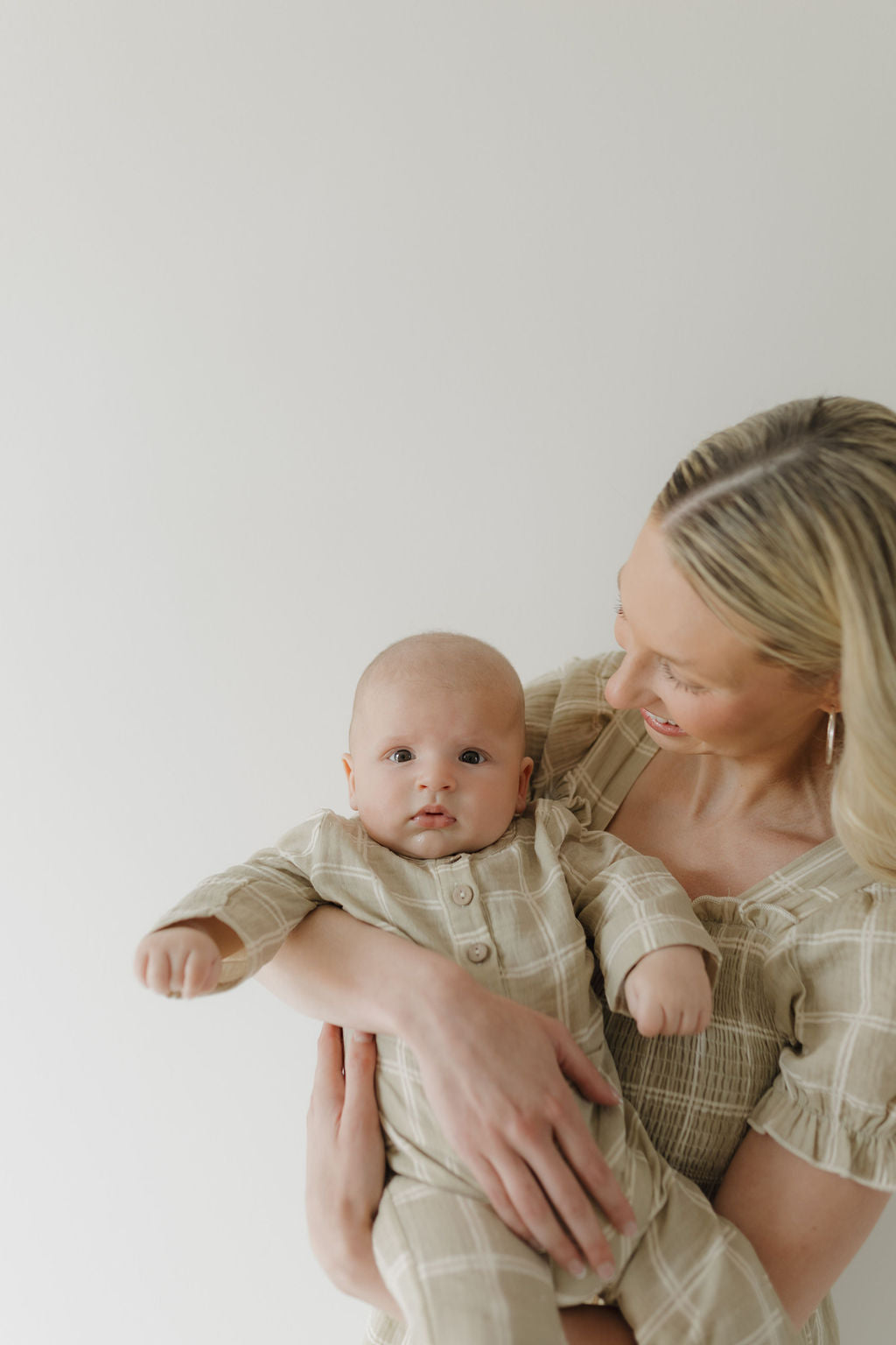 A smiling woman with long blonde hair holds a baby, both dressed in forever french baby's Women's Short Sleeve Muslin Dress | Linen Grid—matching beige plaid outfits crafted from lightweight 100% cotton, set against a light background.