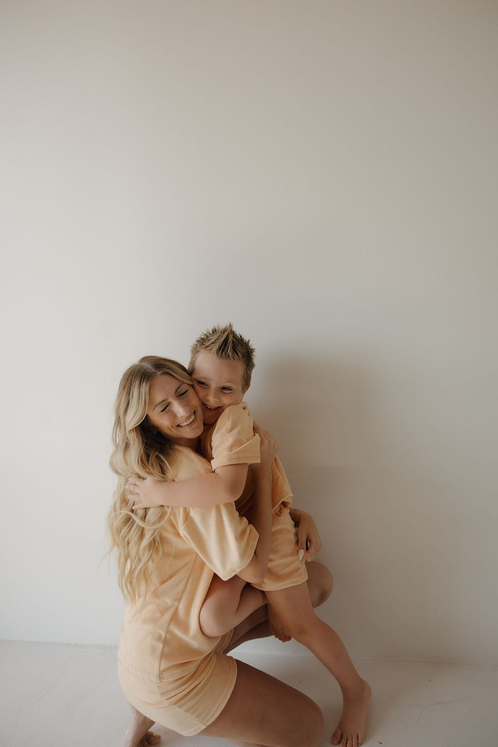 A woman with long blonde hair kneels and hugs a smiling young boy. Both wear matching forever french baby Chid Terry Short Set in Sherbert, capturing sweet family moments against a plain white wall.