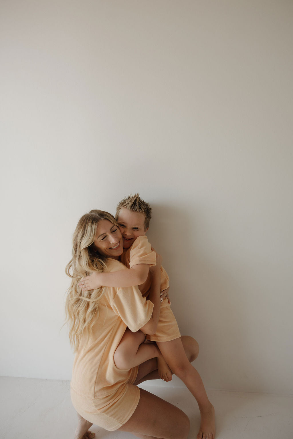 A woman with long blonde hair kneels and hugs a smiling young boy; both wear matching Chid Terry Short Set | Sherbert by forever french baby, set against a simple, light background—capturing a sweet family moment.
