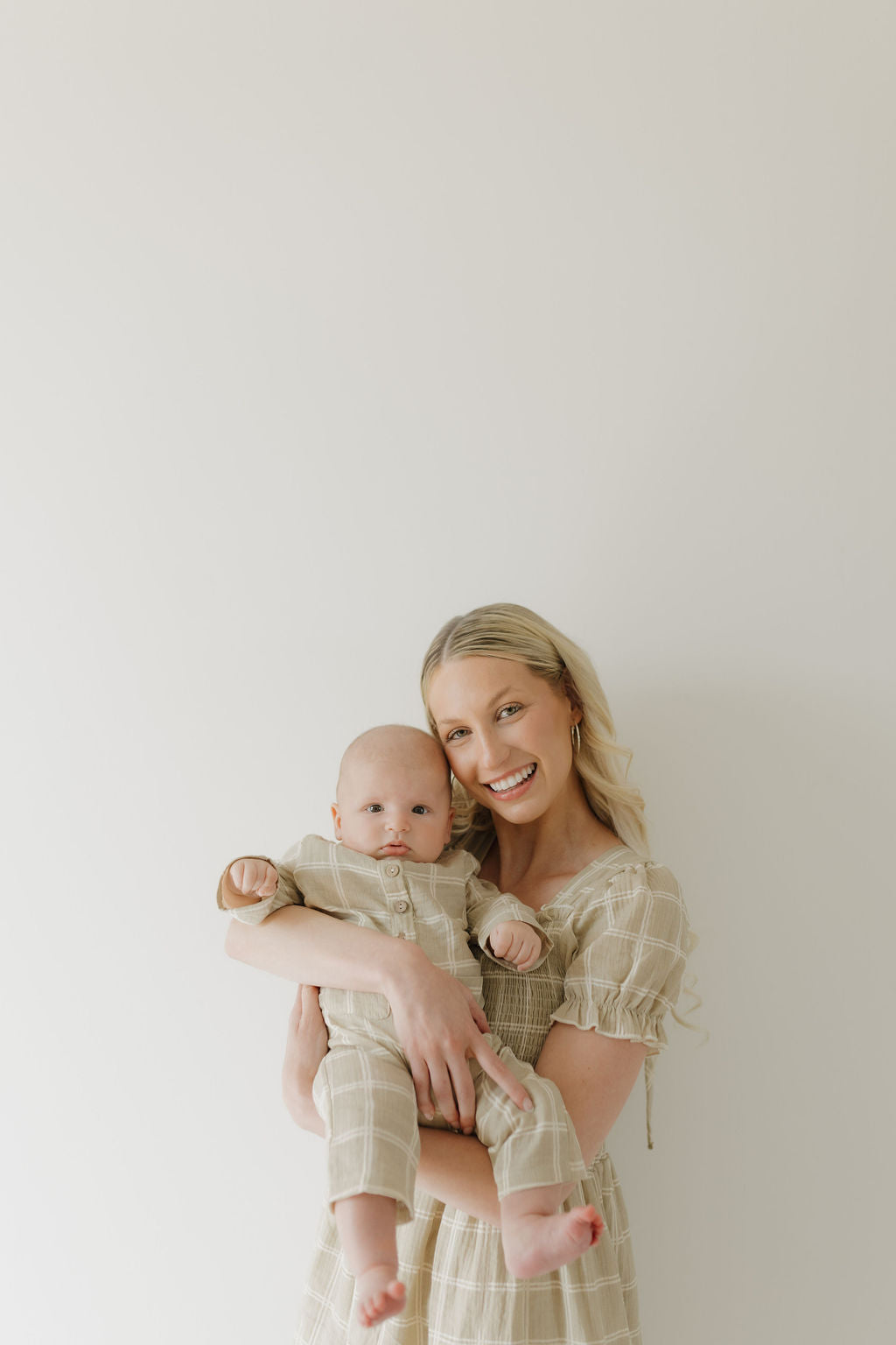 A smiling woman with long blonde hair holds a baby. Both wear matching forever french baby 100% cotton outfits—the woman in the Women's Short Sleeve Muslin Dress | Linen Grid—against a plain light background.