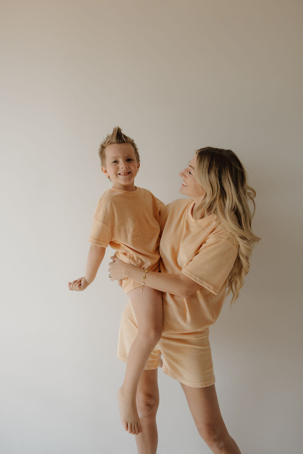 A woman with long blonde hair holds a smiling young boy wearing the forever french baby Chid Terry Short Set in Sherbert, capturing sweet family moments against a plain, light background.