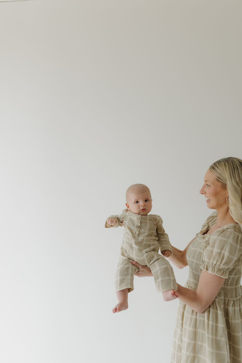 A woman in a forever french baby Women's Short Sleeve Muslin Dress | Linen Grid, with long blonde hair, smiles warmly at a baby in beige plaid. Both face left against a plain light background.