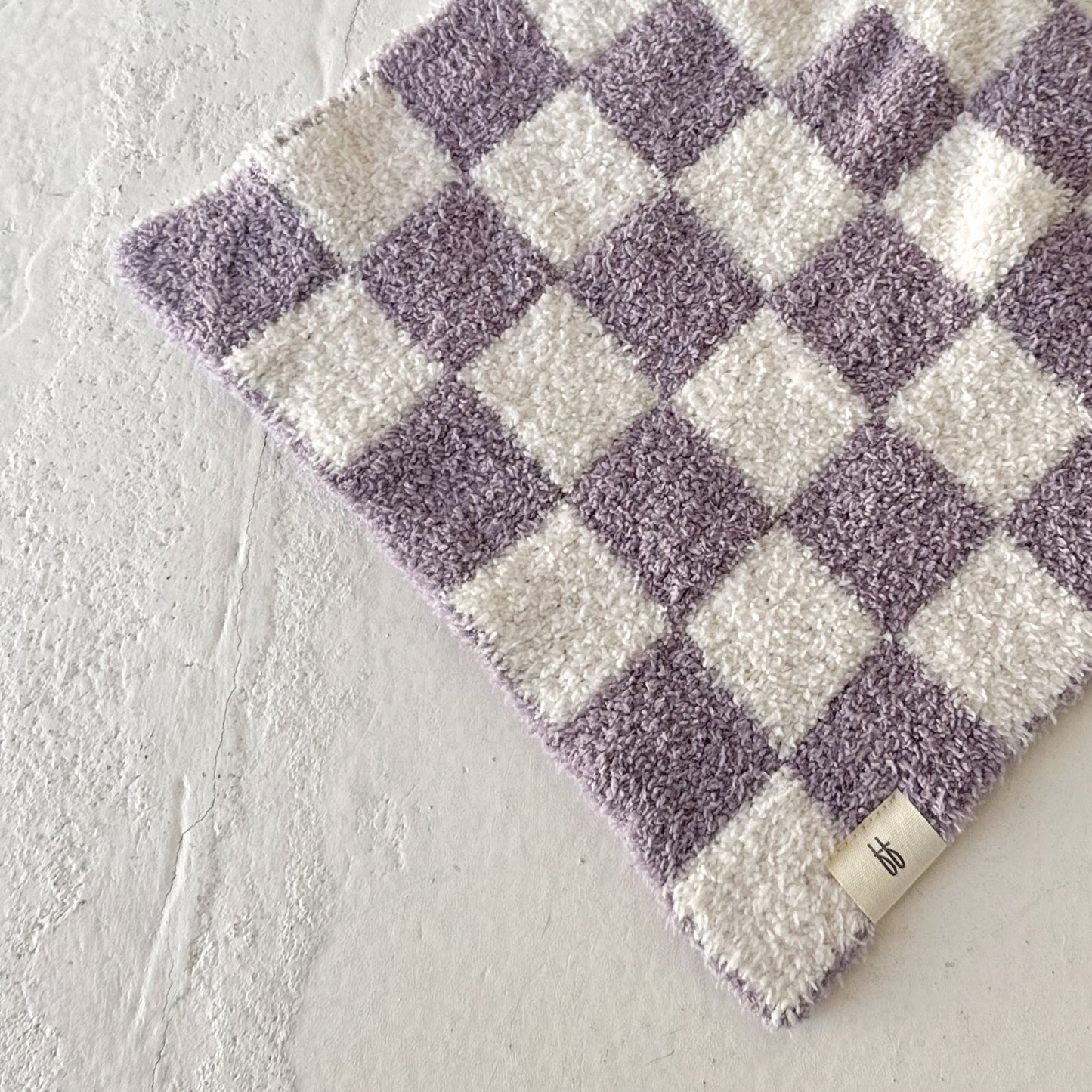 The Lovey | Grape Soda by forever french baby is a soft, microfiber baby blanket in light purple and white checkers, shown partially folded with a beige branded tag on one corner.