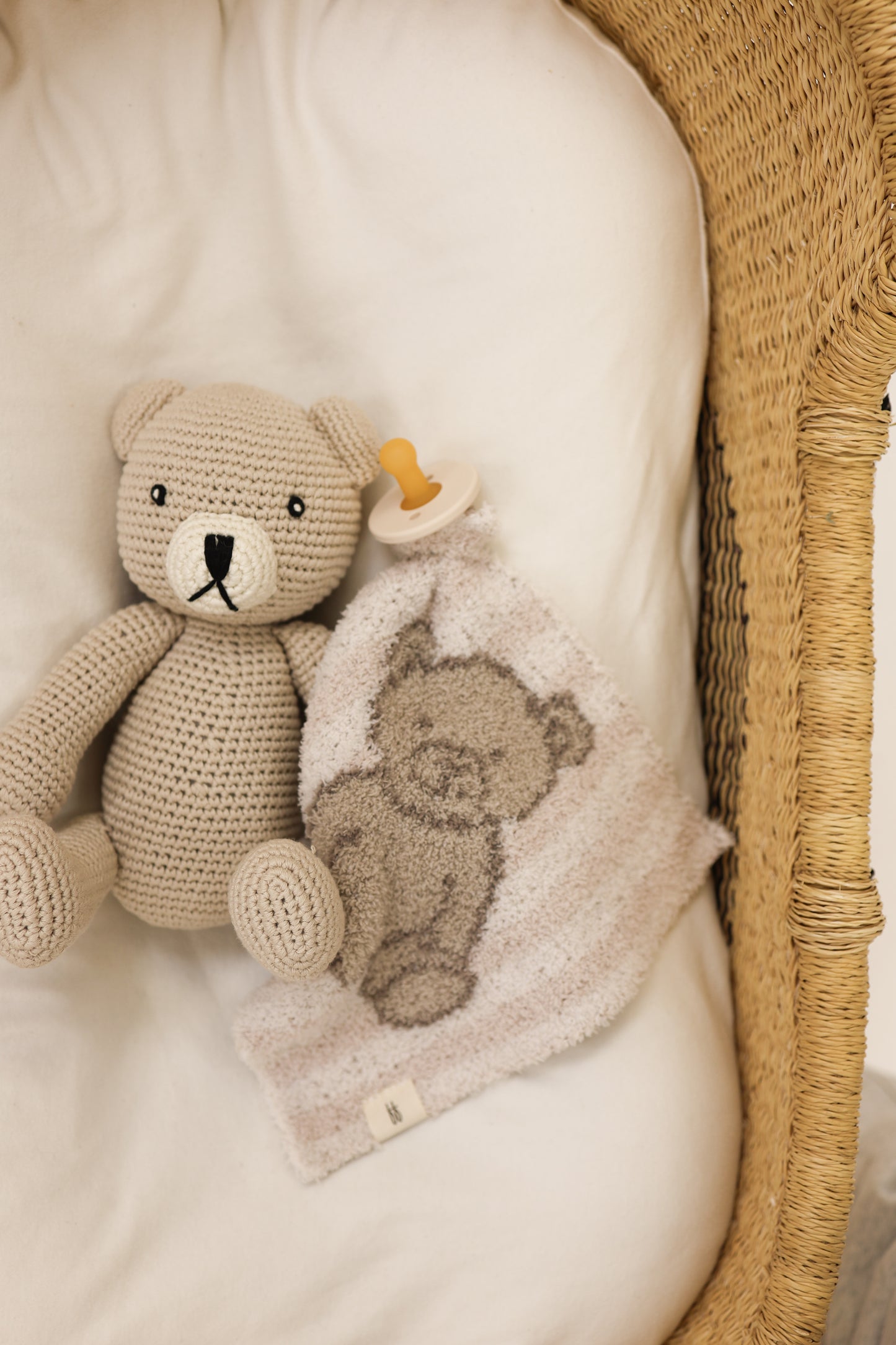 The Lovey | Teddy by forever french baby—a crocheted beige teddy bear, beige pacifier, and plush microfiber bear blanket—rests on a white sheet inside a woven basket bassinet.